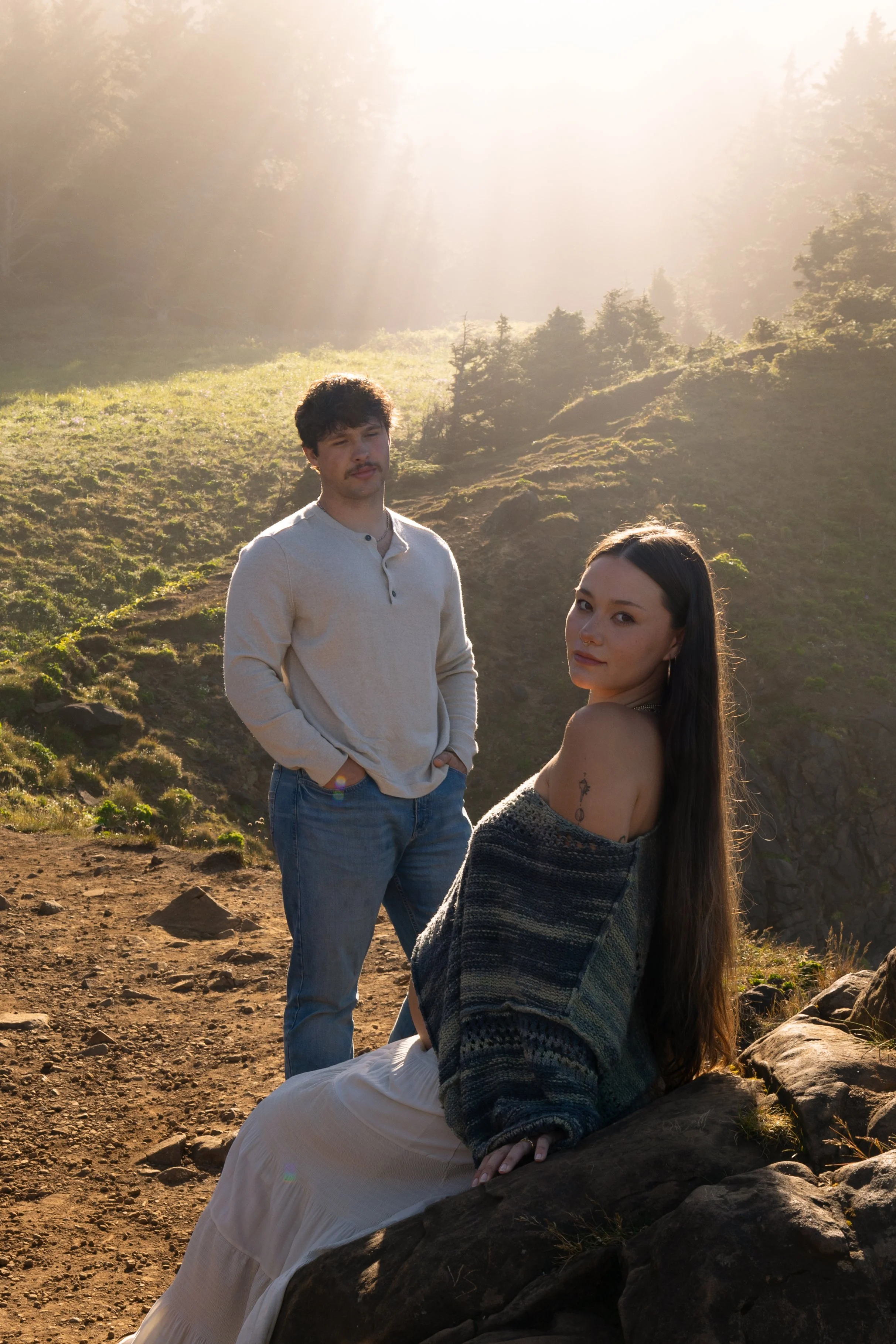 A man and woman outdoors on a hillside during golden hour sunlight, the woman sitting on a large rock and the man standing with hands in pockets, both looking toward the camera.