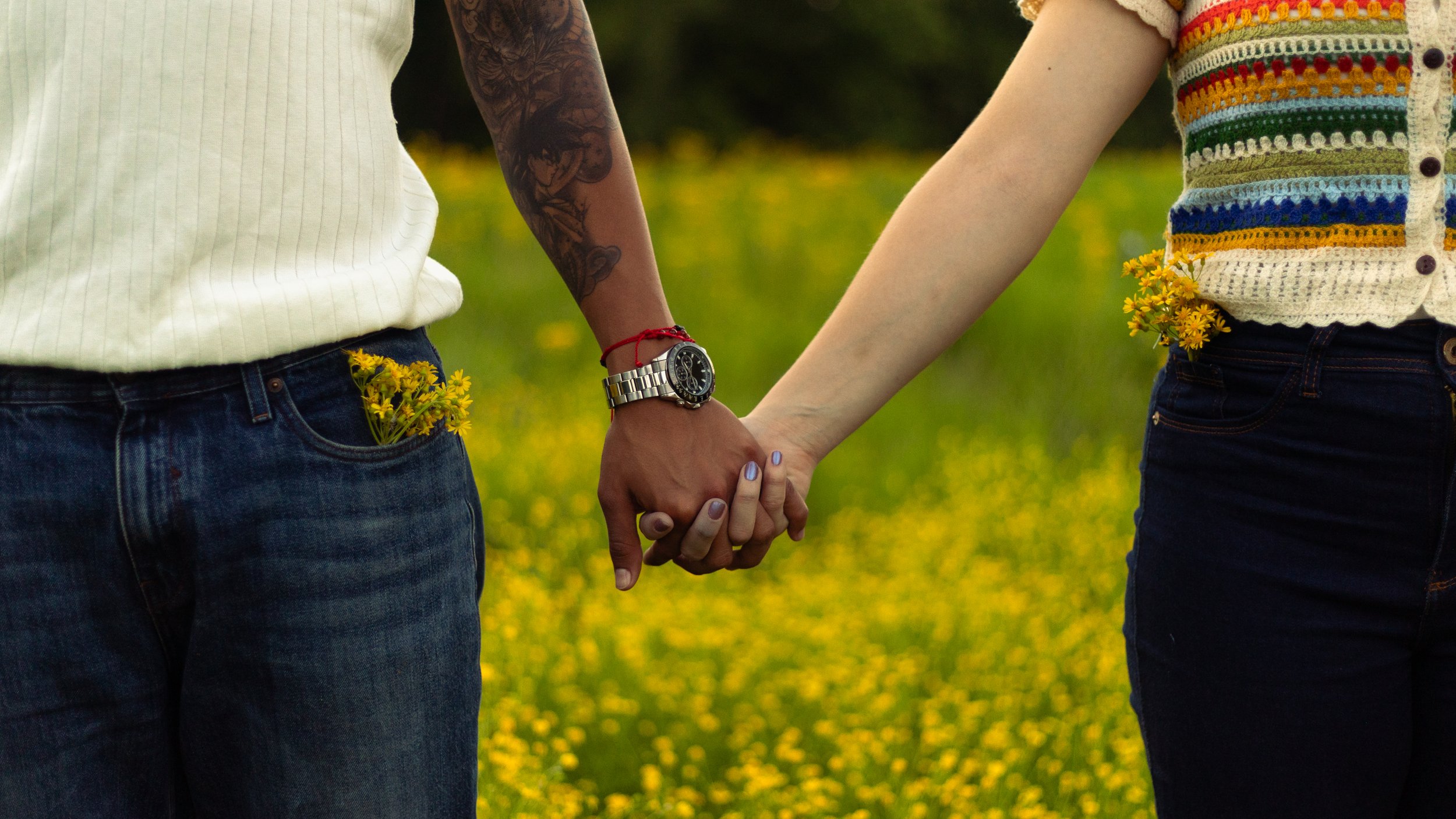 A close-up of a couple holding hands in a field of yellow flowers, with their torsos visible. One person wears a white tank top and a watch, while the other wears a colorful crocheted top.