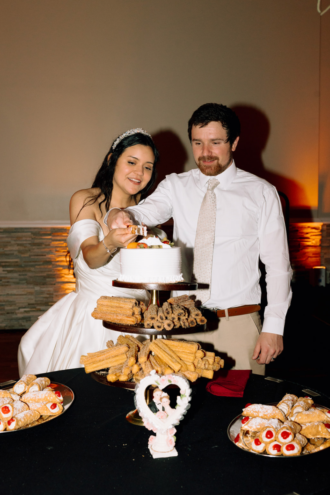 A newlywed couple cutting their wedding cake at a reception. The bride is in a white dress with an off-shoulder design and a tiara, and the groom is in a white shirt with a light-colored tie. They are standing behind a multi-tiered tray filled with c