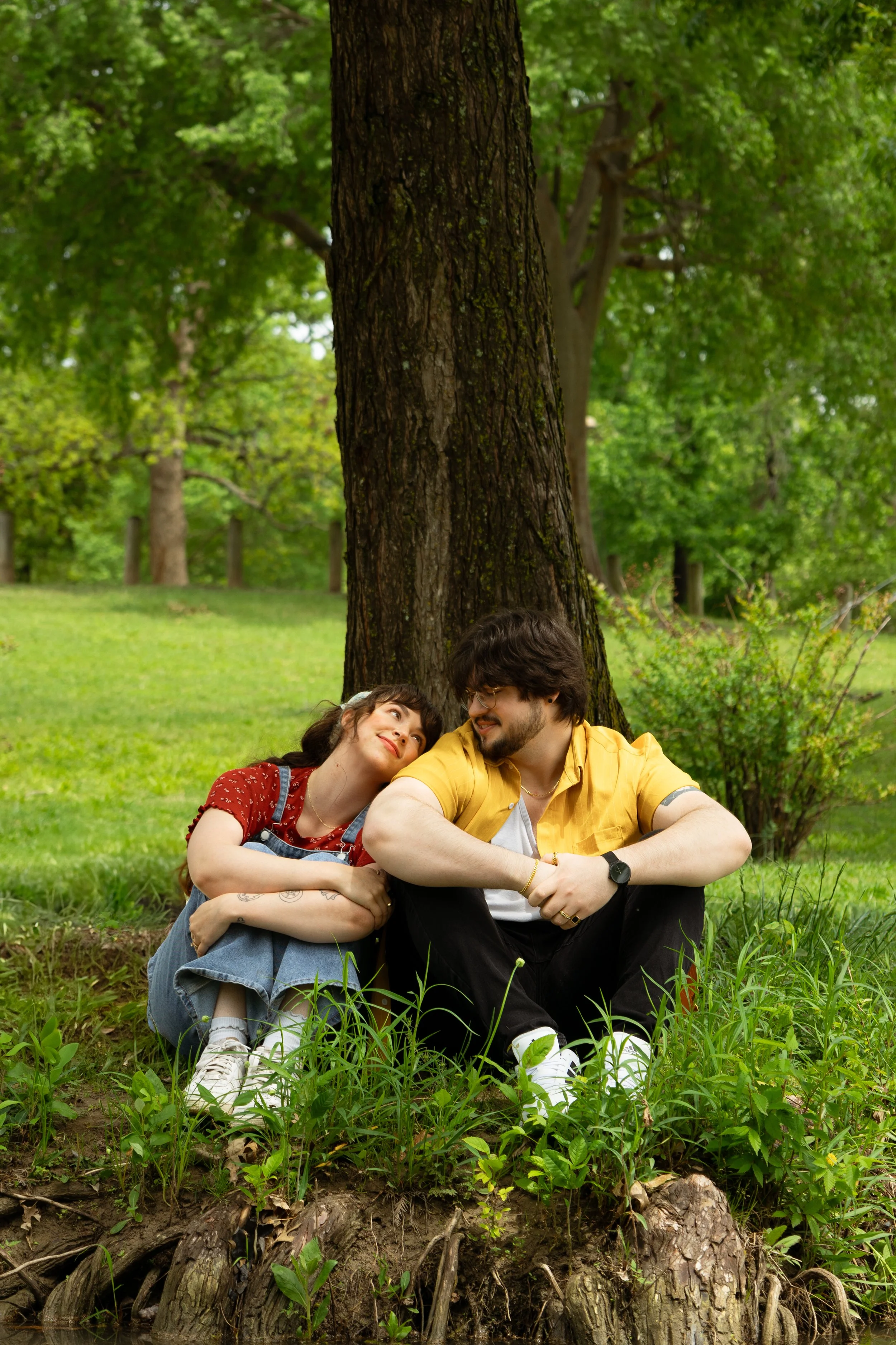 A young couple sitting under a large tree on grass in a park, smiling and looking at each other.