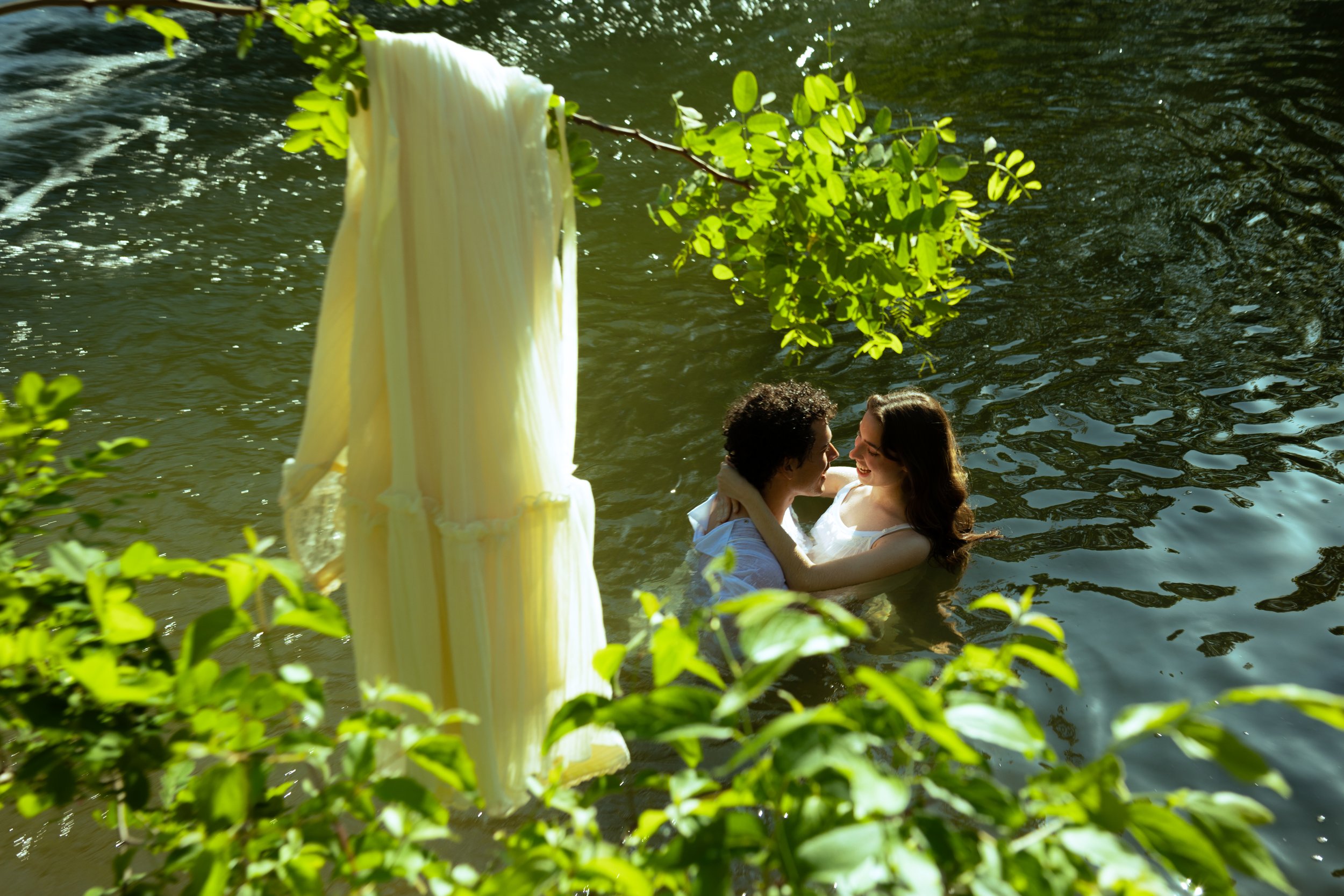A couple wading in a river, holding each other and smiling, with a yellow dress hanging on a branch above them.