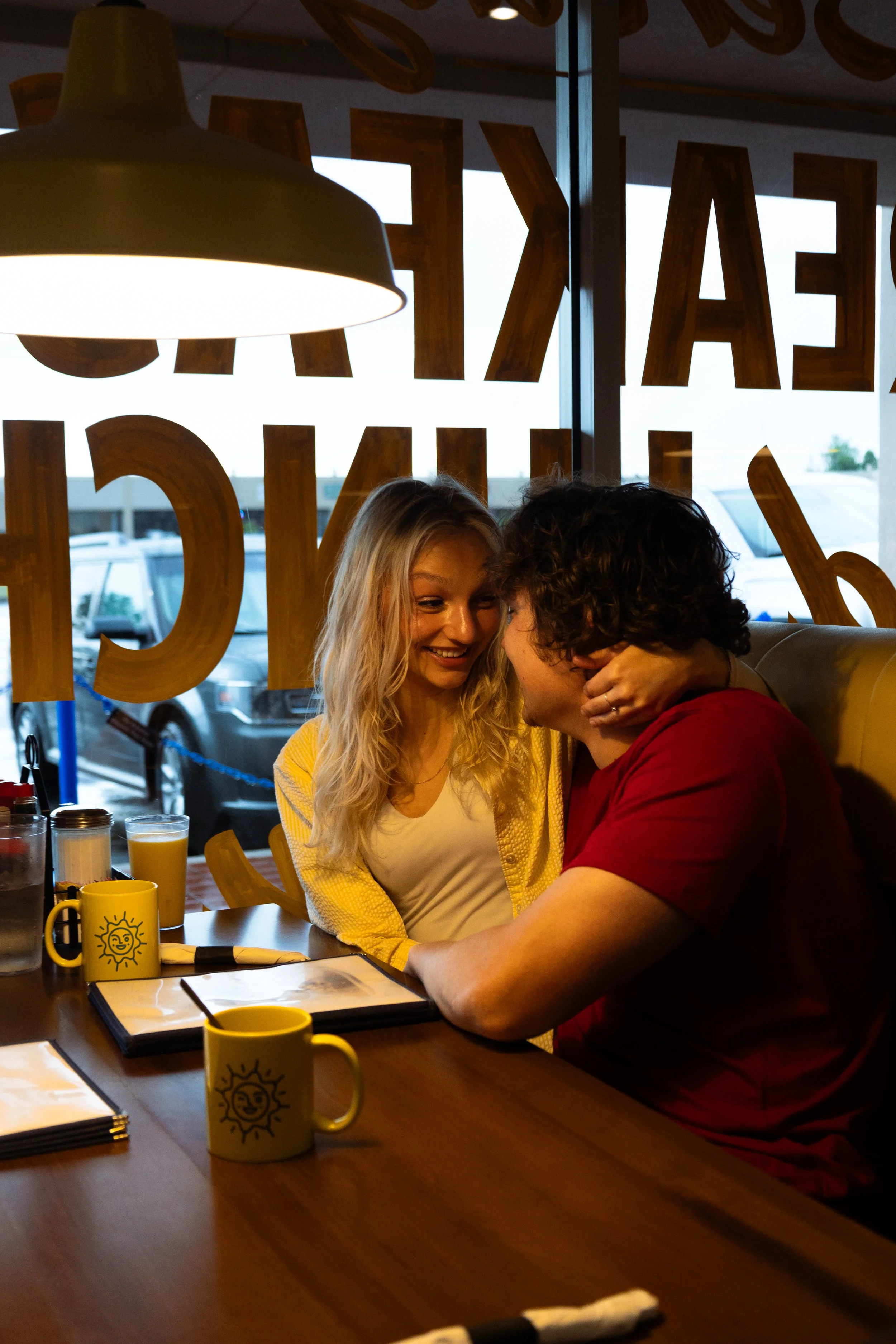 A man and woman sharing a close moment at a restaurant table, with drinks and menus in front of them, sitting by a window with large letters on the glass.