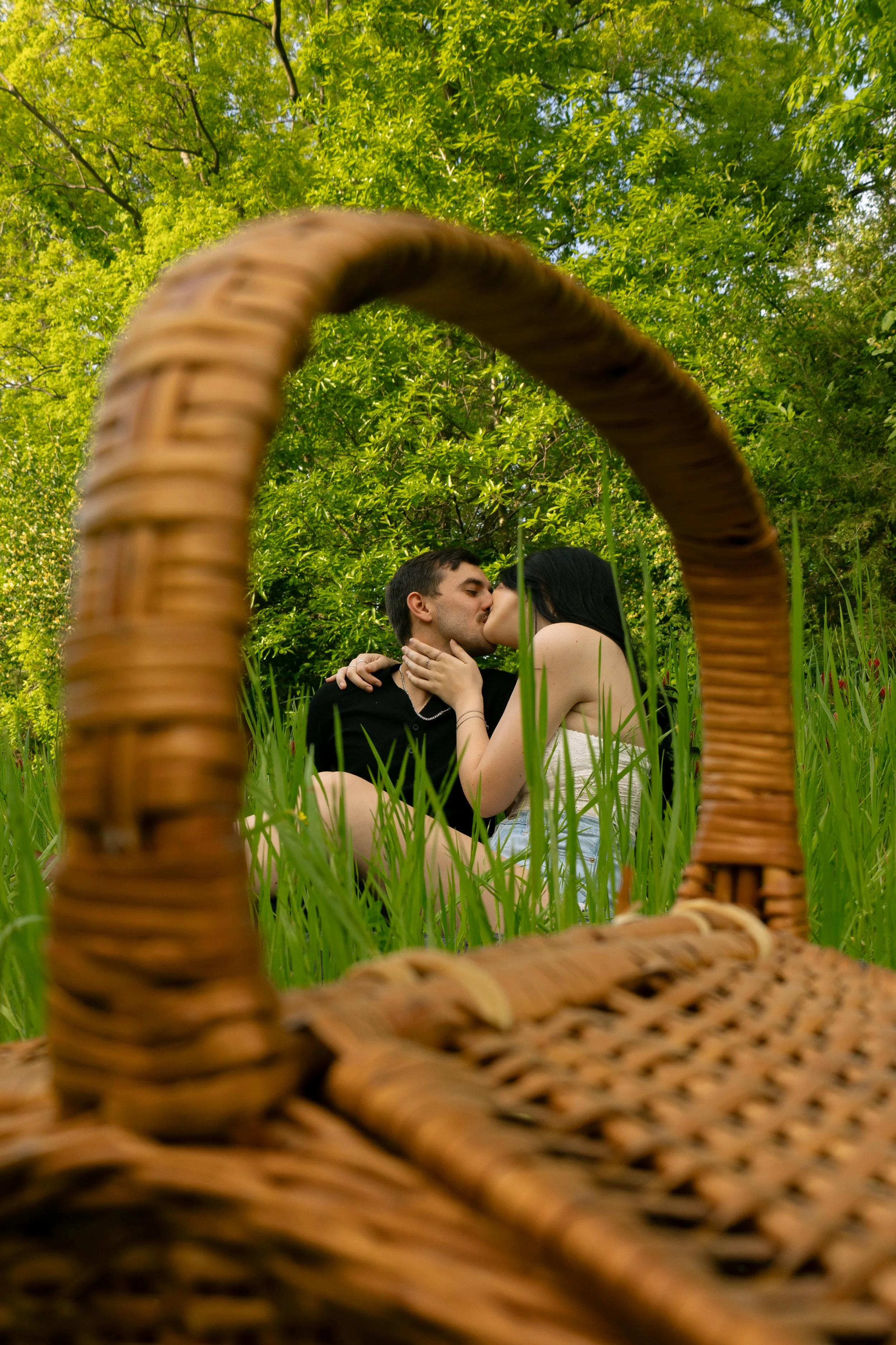 A couple kissing in a grassy, wooded outdoor setting seen through a woven wicker basket.