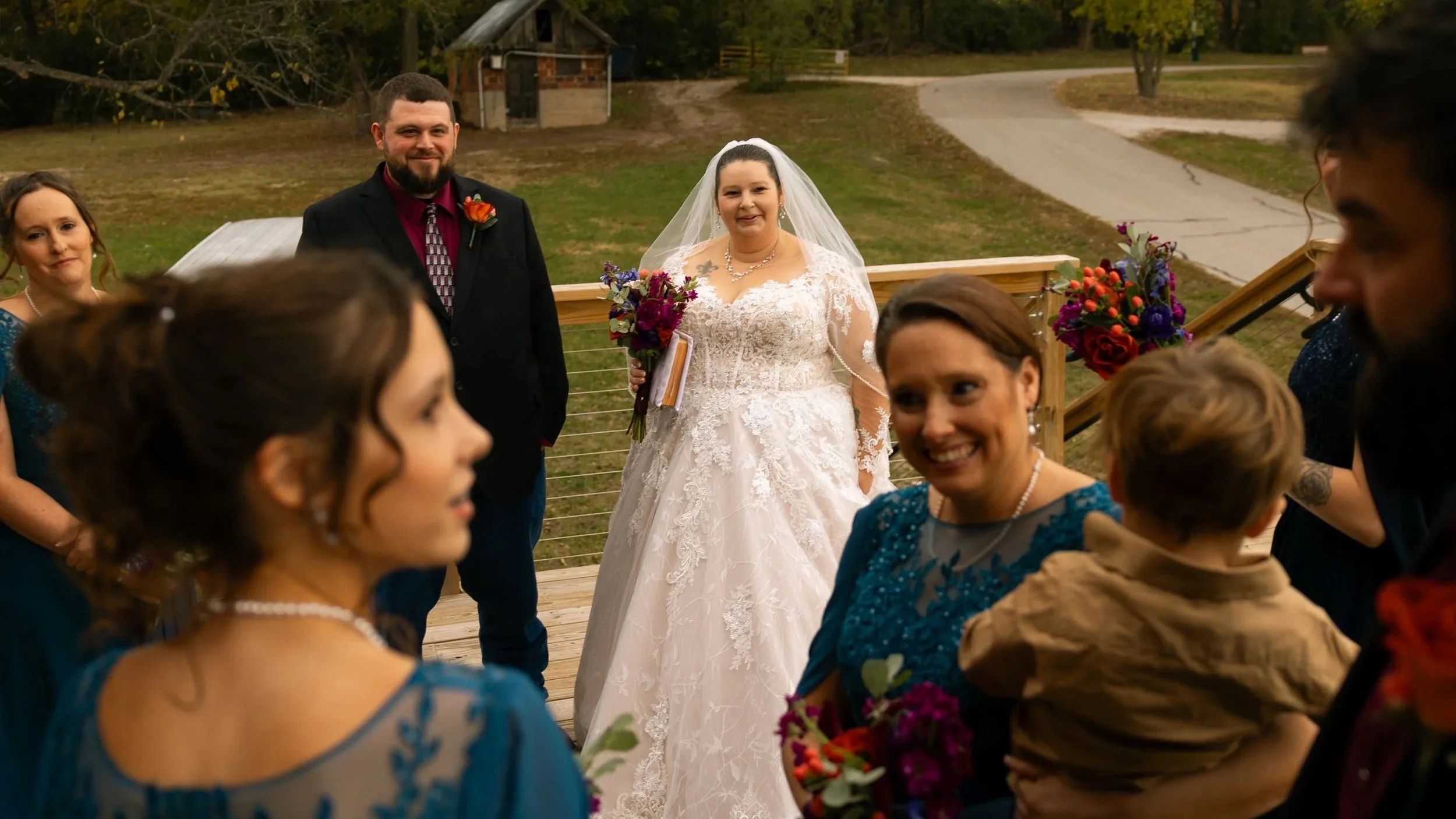 Bride in a white lace wedding gown and veil holding a bouquet, smiling, standing outdoors on a wooden deck with guests gathered around, during a wedding celebration.
