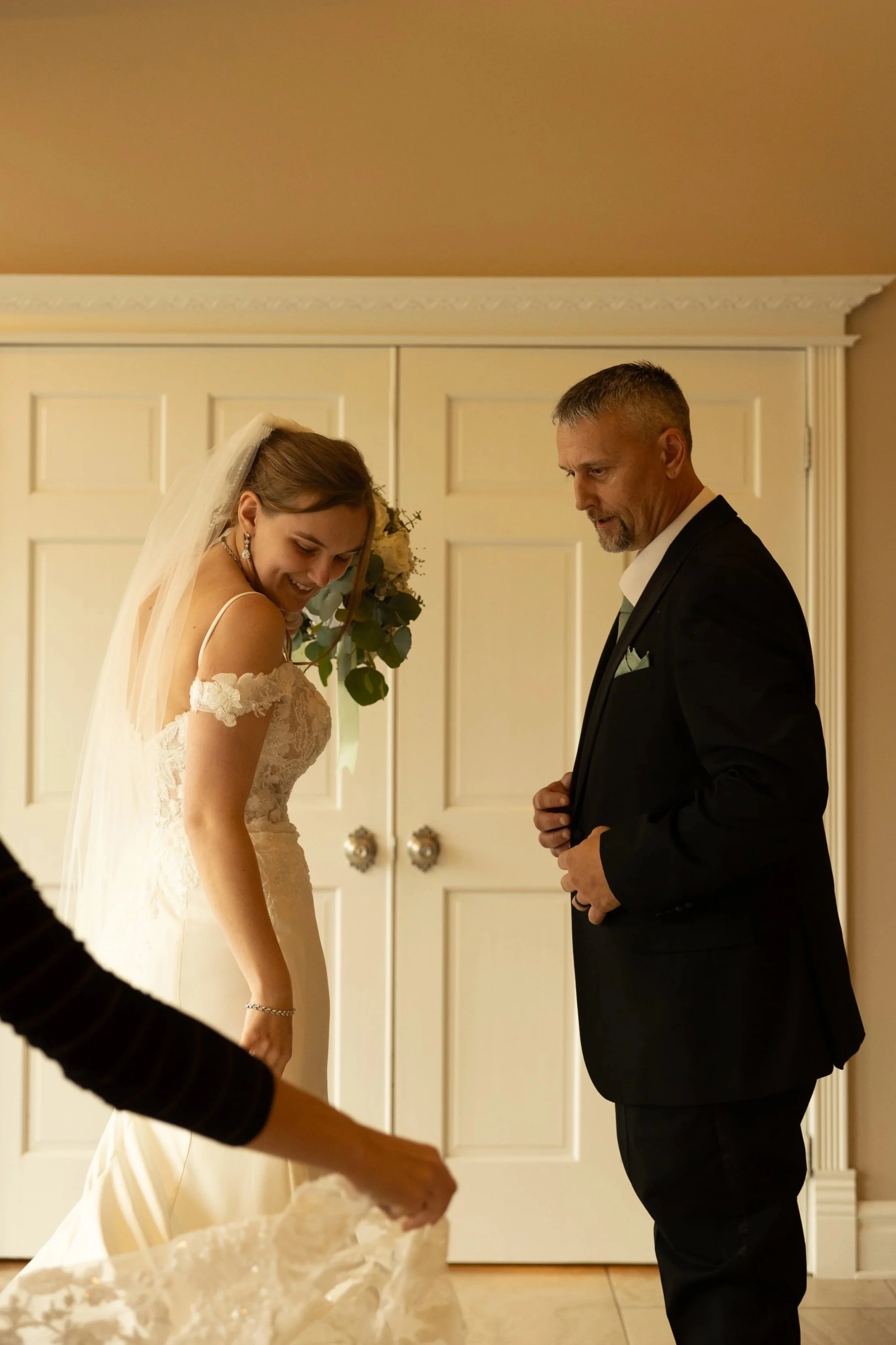 A bride in a wedding dress smiling and holding a bouquet, standing in front of an older man in a suit, indoors near a closed white door.
