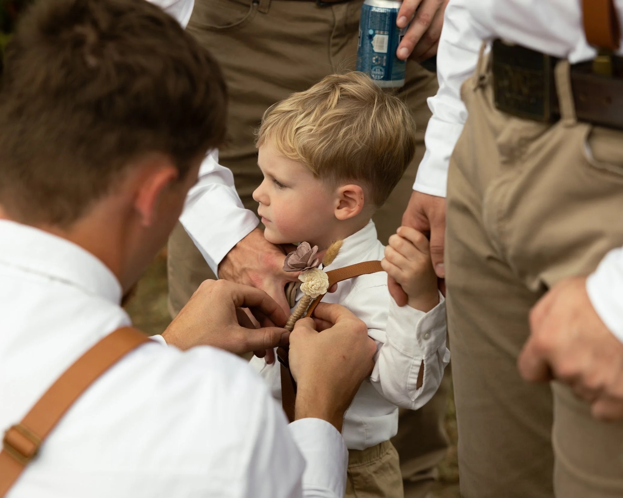 A young boy is being dressed with a bowtie by an adult, while other adults surround him, in an outdoor setting.