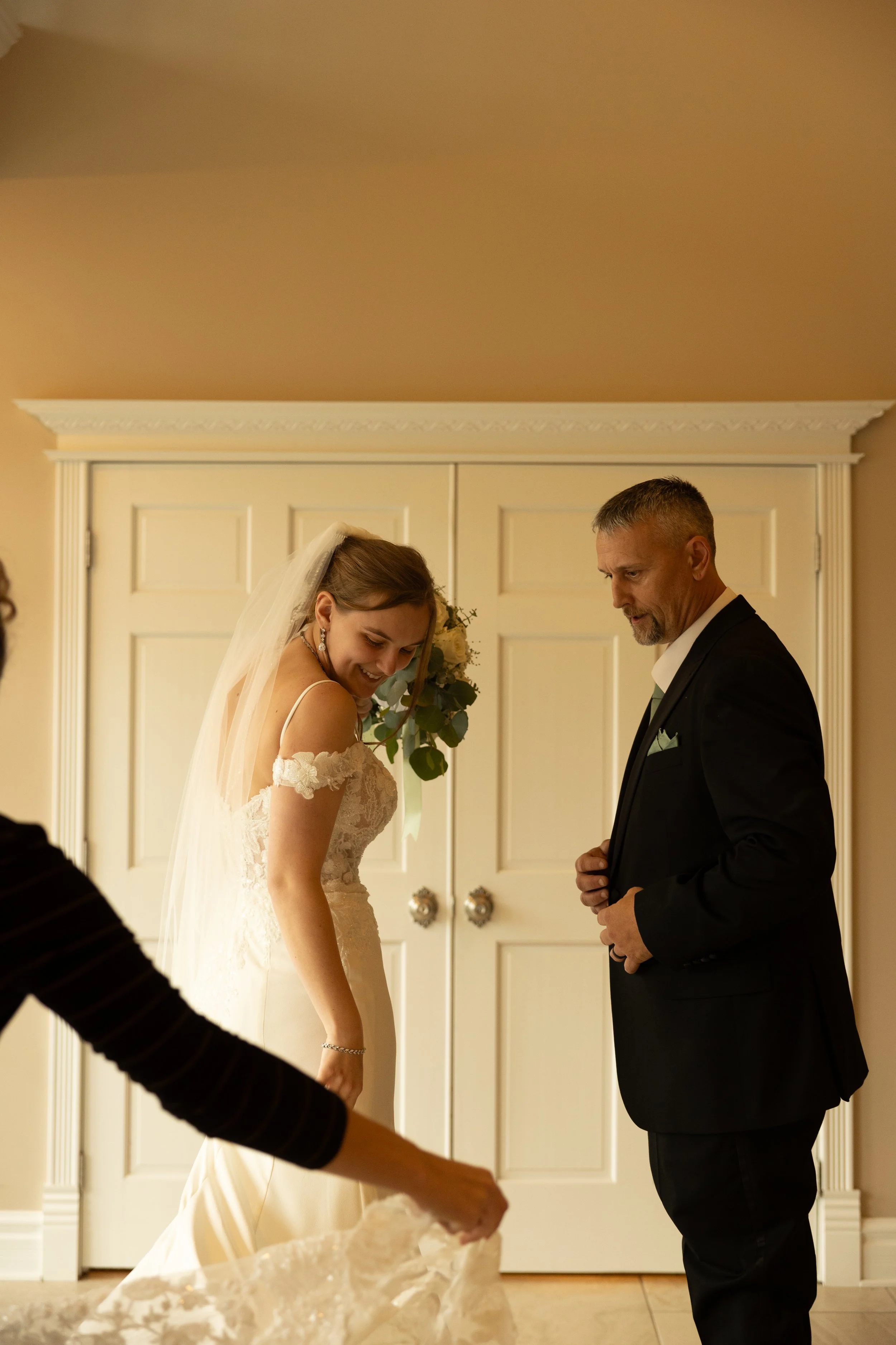 Bride in wedding dress smiling at her father on her wedding day in a warmly lit room with cream-colored walls and closed double doors.