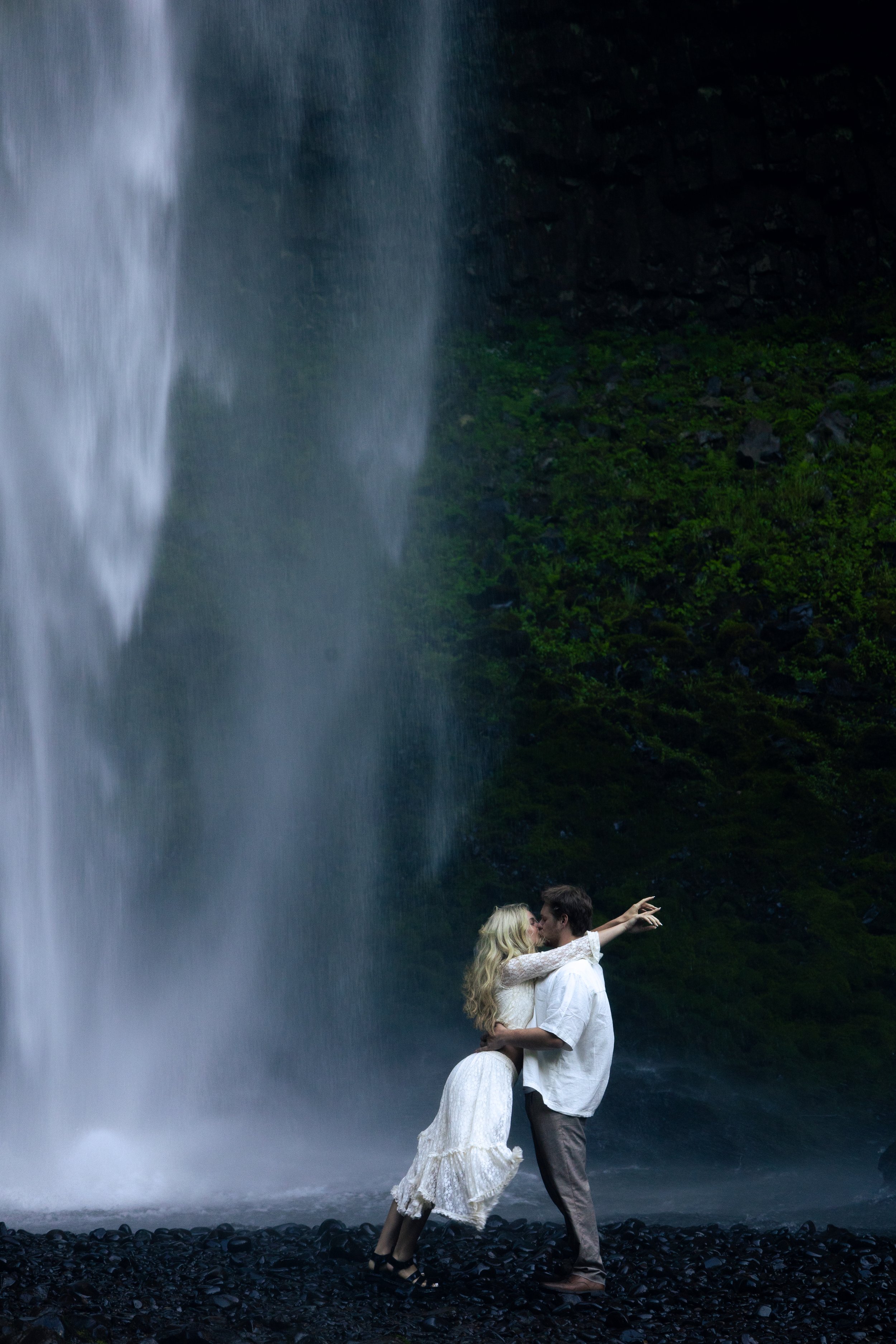 A couple kissing near a large waterfall with a green mossy cliff in the background.