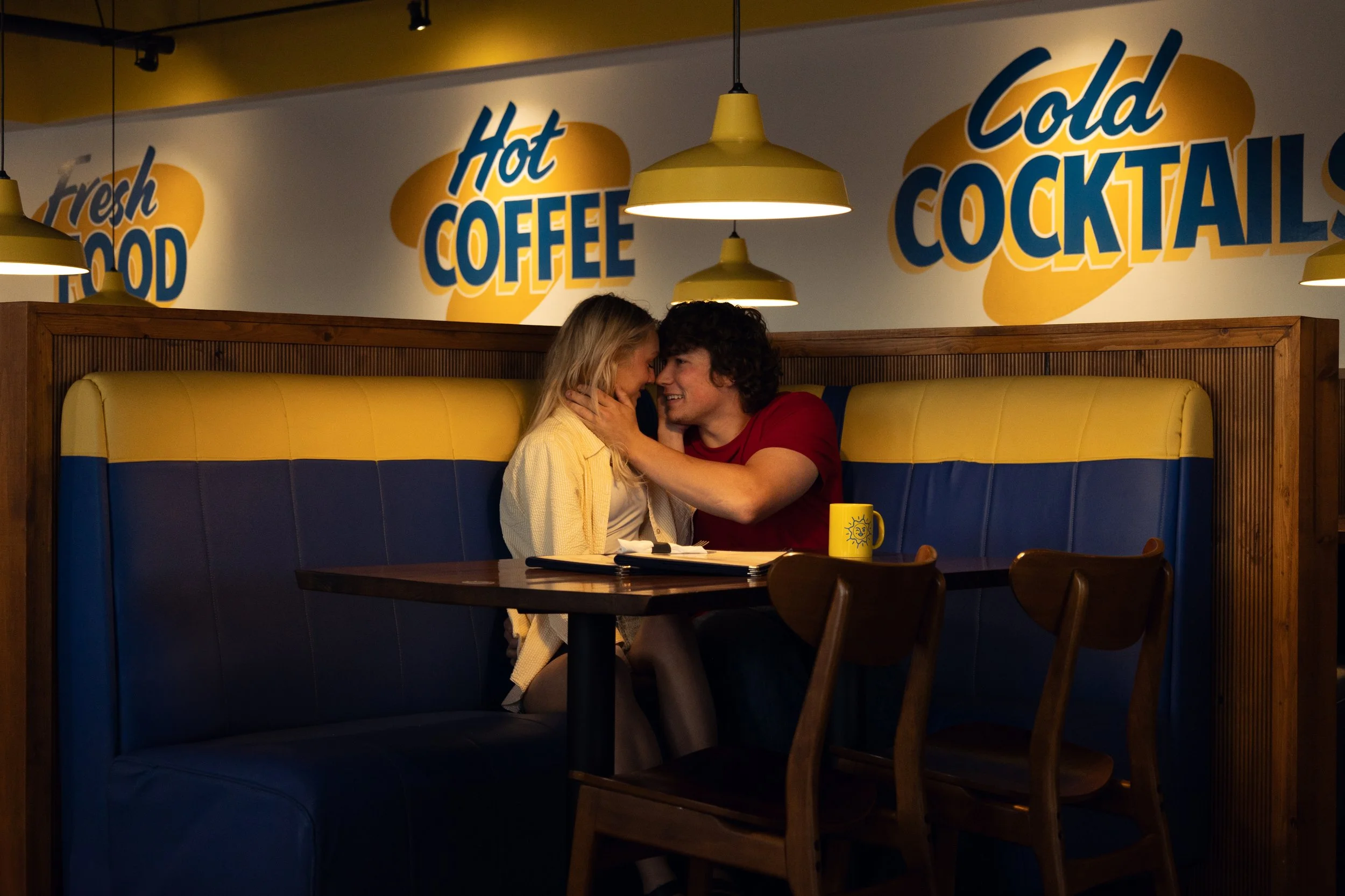 A couple sitting closely in a booth at a restaurant, with the man touching the woman's face, surrounded by signs that say 'Fresh Food,' 'Hot Coffee,' and 'Cold Cocktails'.