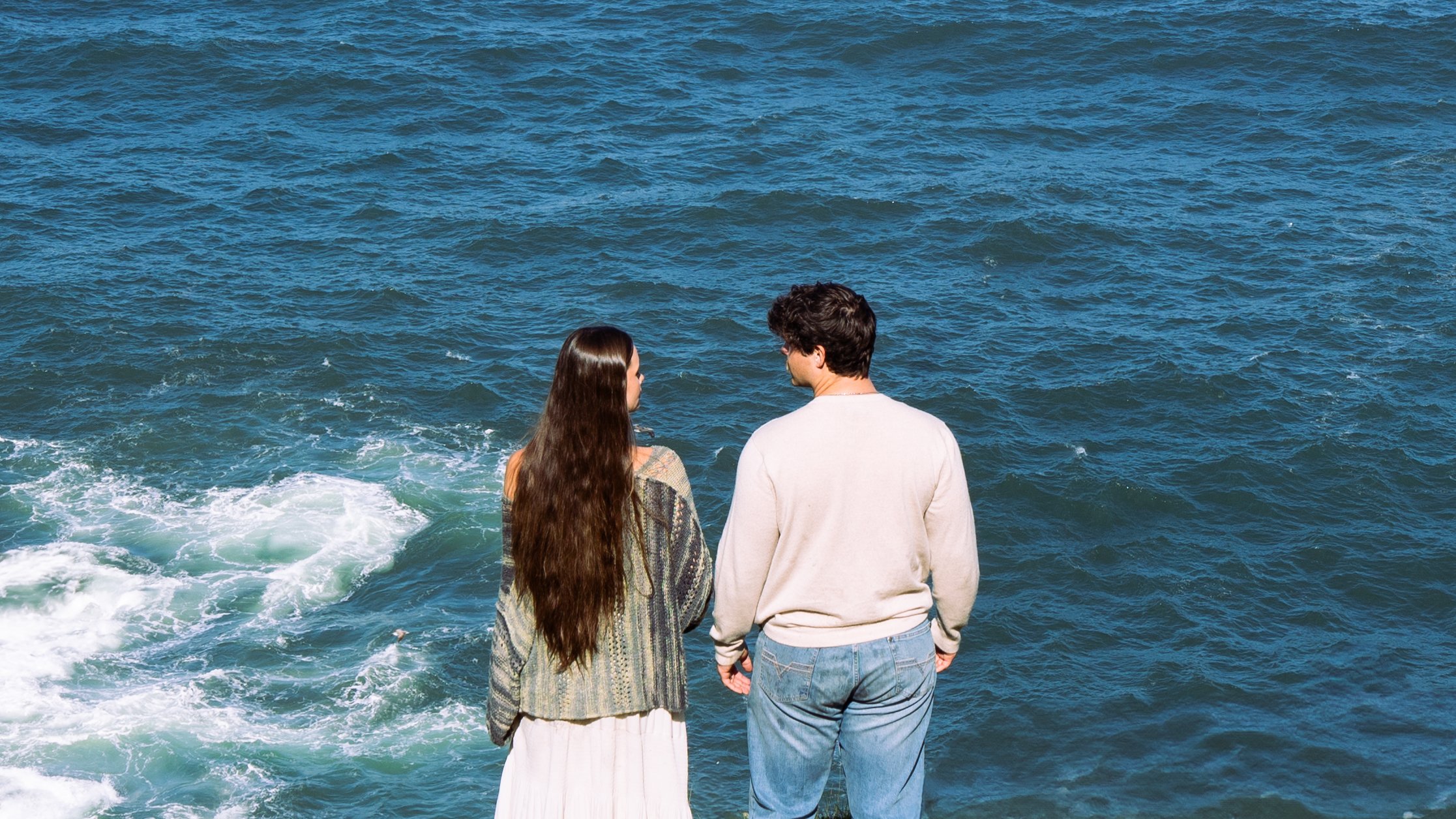 A man and woman stand by the ocean, talking to each other.