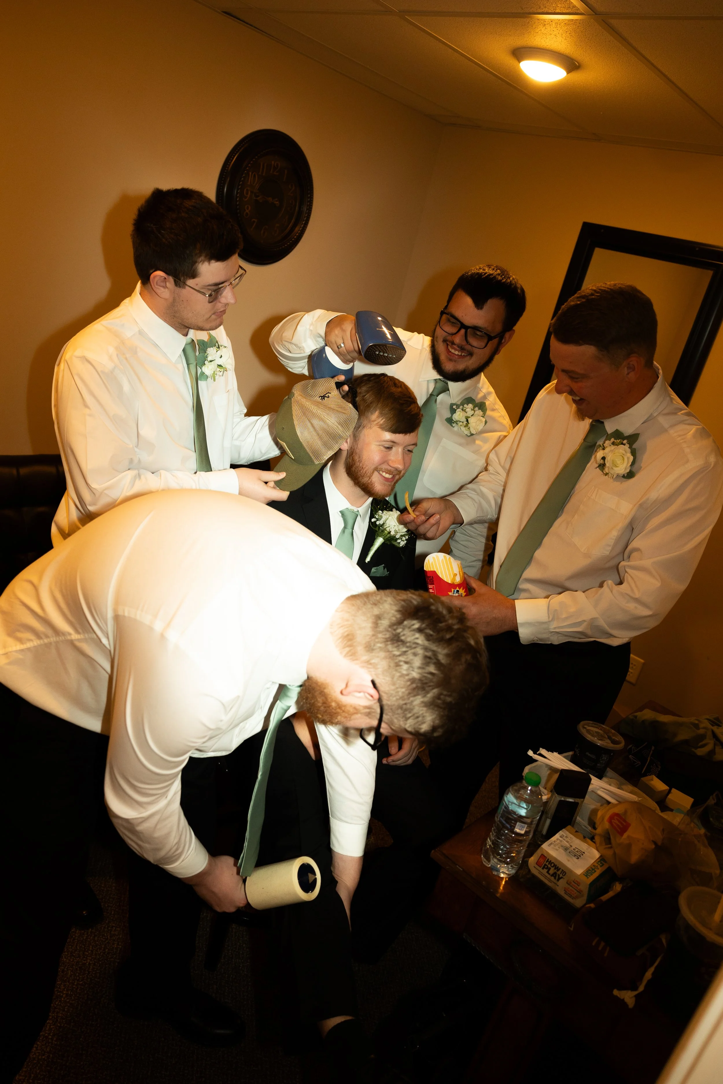 Group of men, some with glasses, helping groom with hairstyle and hairdryer, in indoor wedding setting with beige walls, clock, and mirror.
