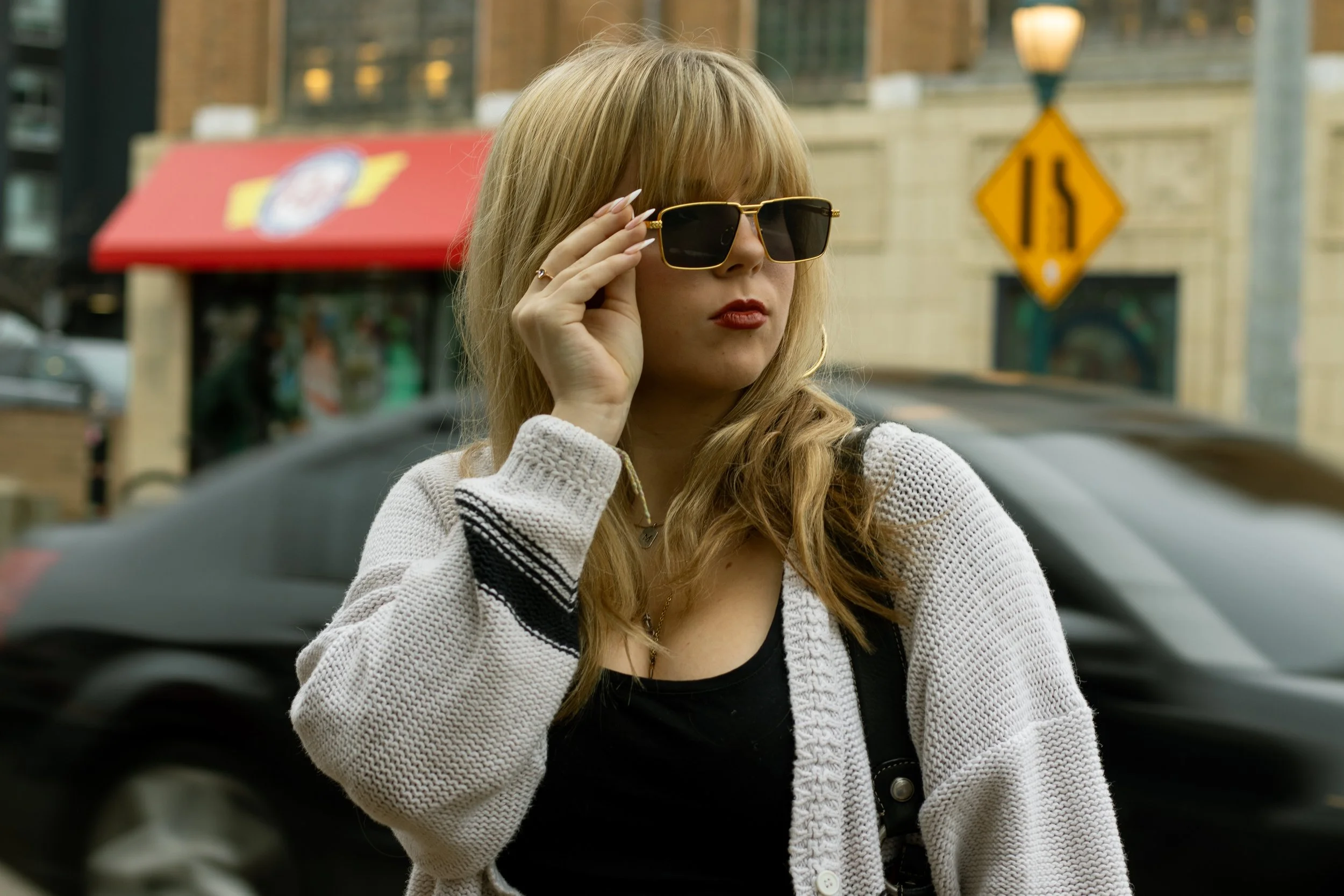 A woman with long blonde hair wearing sunglasses, a black top, and a white cardigan, standing on a city street with cars and storefronts in the background.