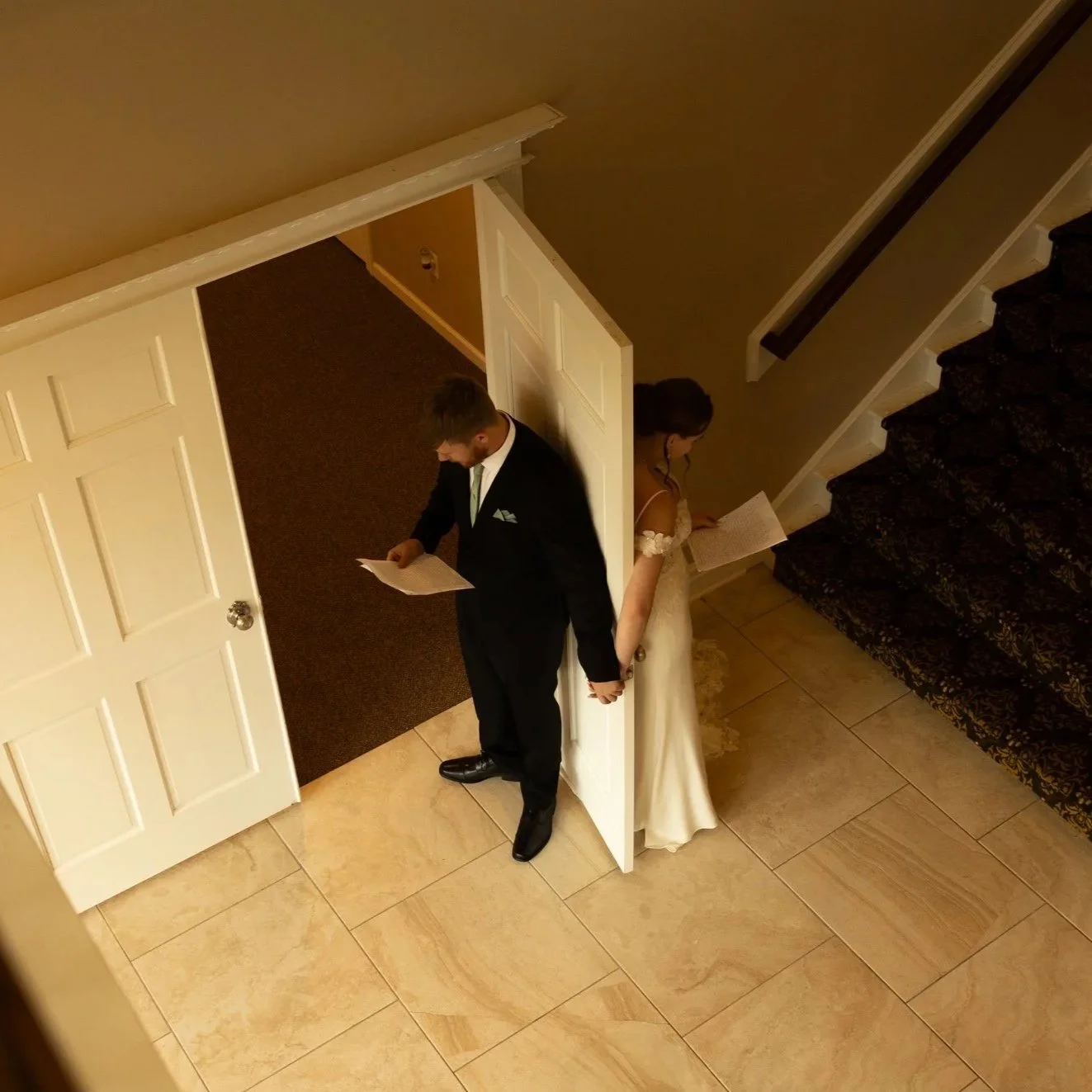 A bride and groom standing back to back while holding hands, reading papers, with a door and staircase nearby, in a wedding setting.