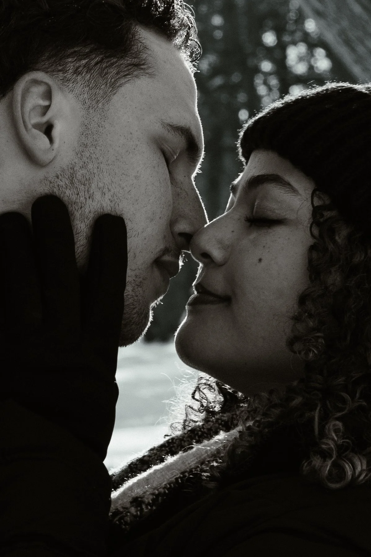 A black and white photo of a man and woman close to each other with eyes closed, almost touching noses, with a natural outdoor background.