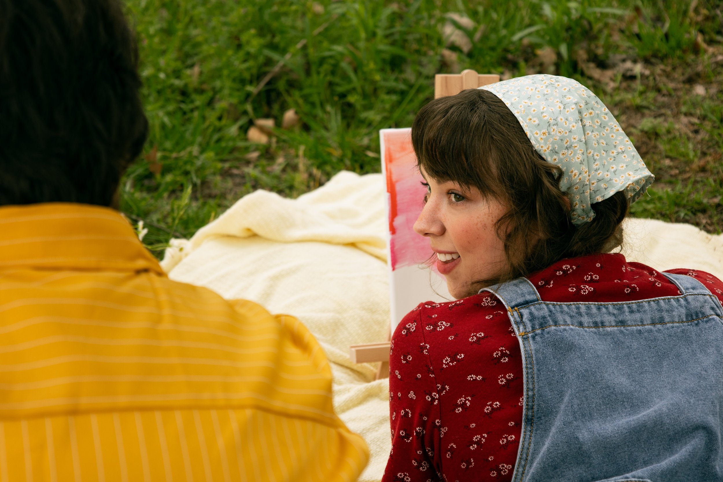 A young woman with a bandana on her head, wearing a red floral shirt and denim overalls, sitting outdoors on a blanket and smiling at another person. Green grass and a small painting on an easel are in the background.