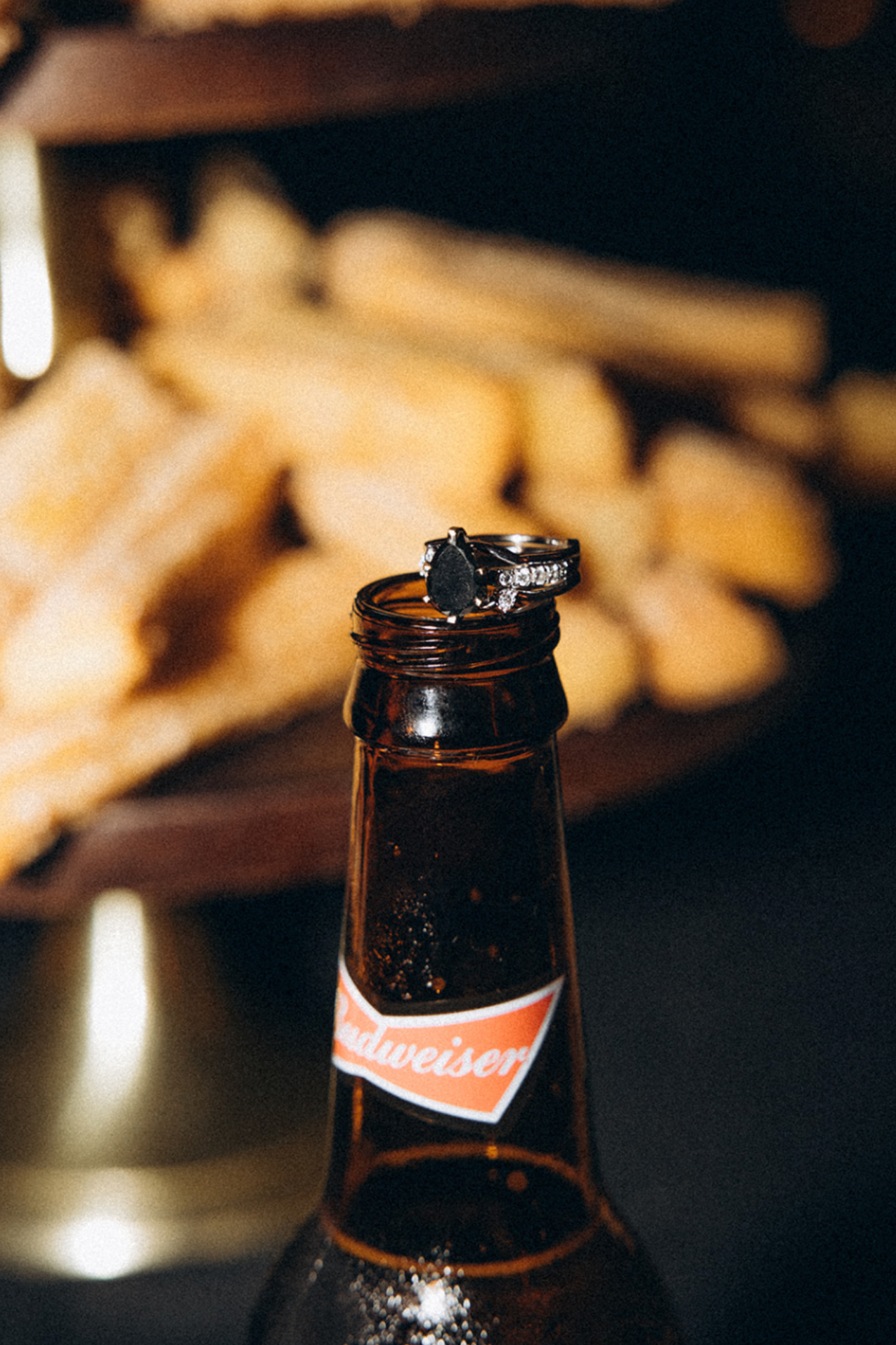 A close-up of a brown glass Budweiser beer bottle with an engagement ring resting on top of its open mouth, with a blurred background of breadsticks and a dark scene.