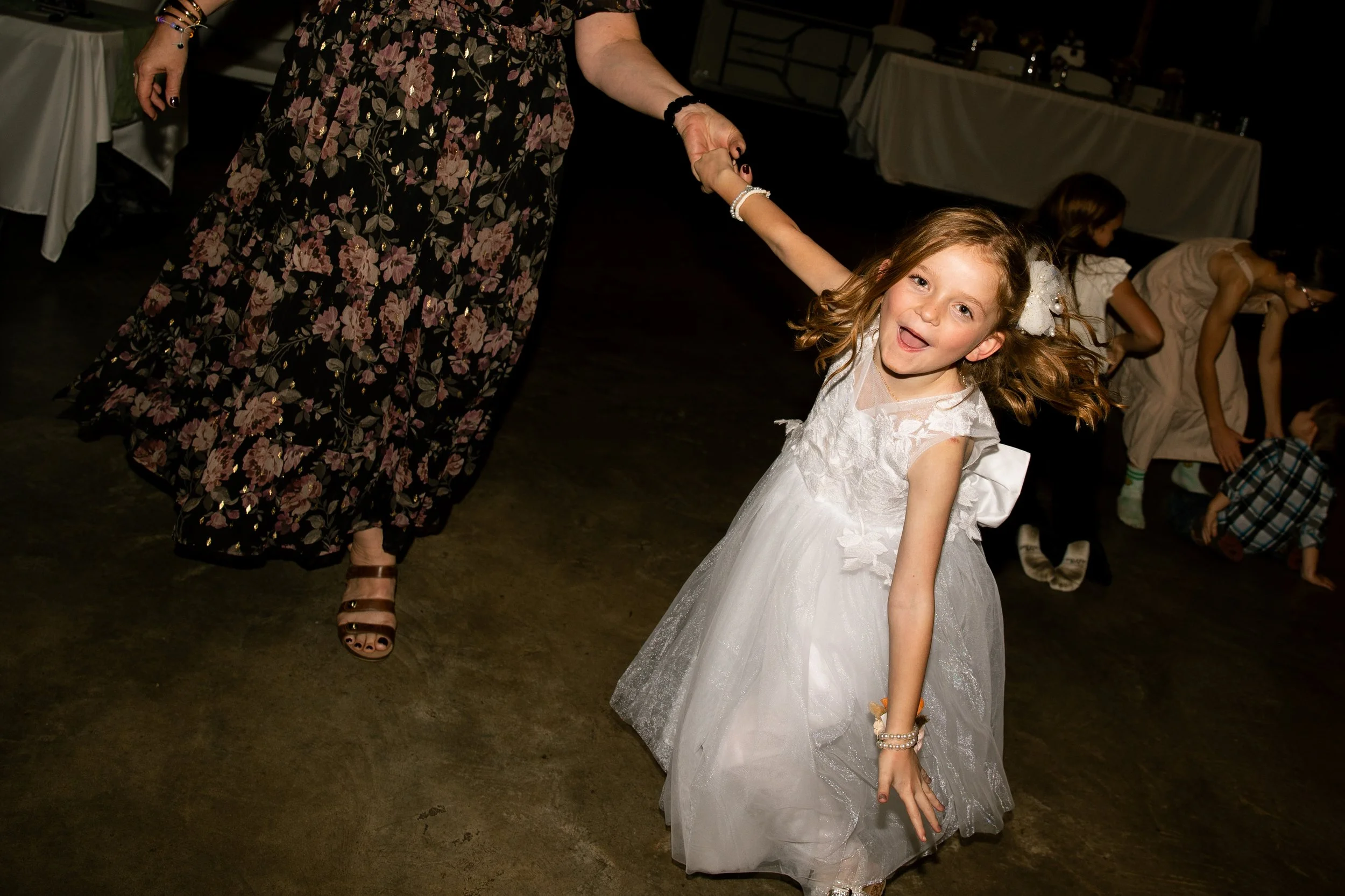 A young girl in a white dress dancing and smiling at a party, holding hands with an adult woman.