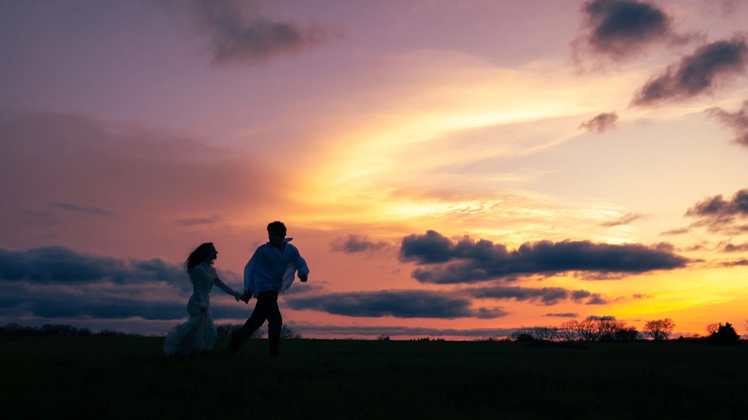 A silhouette of a couple holding hands and running in an open field during sunset, with colorful sky and scattered clouds.