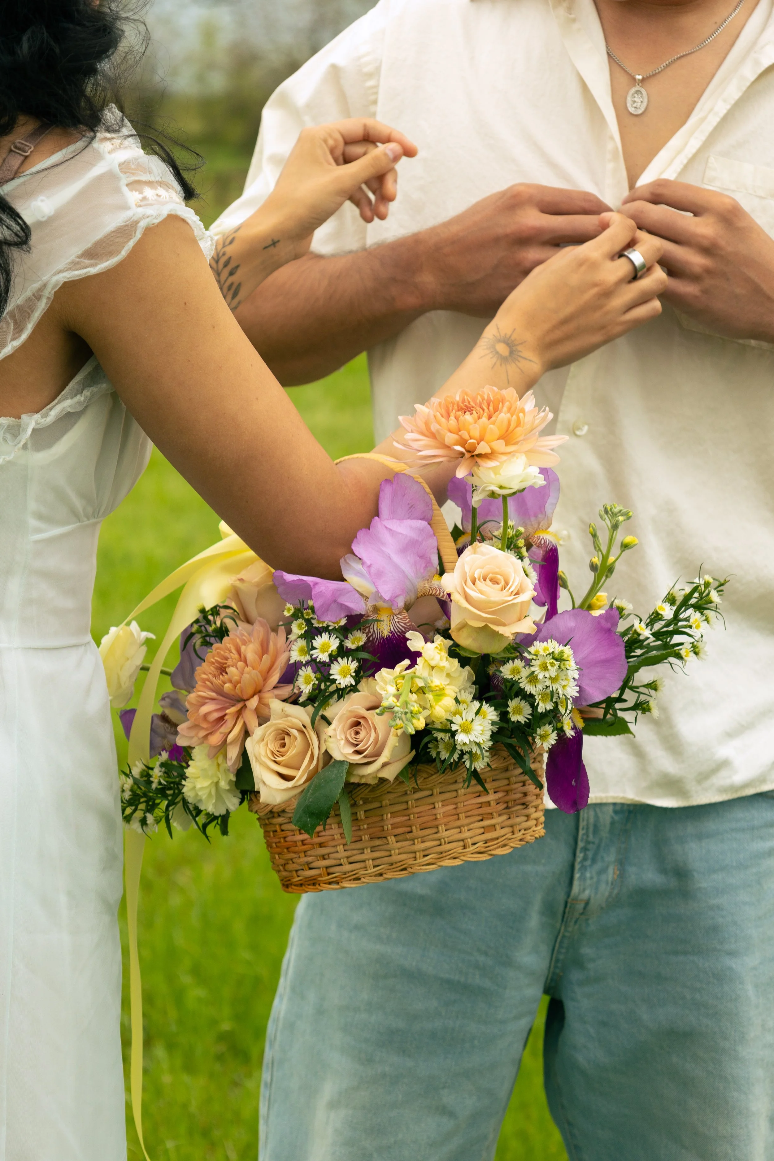 Close-up of two people, one putting a ring on the other's finger, with a basket of colorful flowers in the foreground, outdoors.