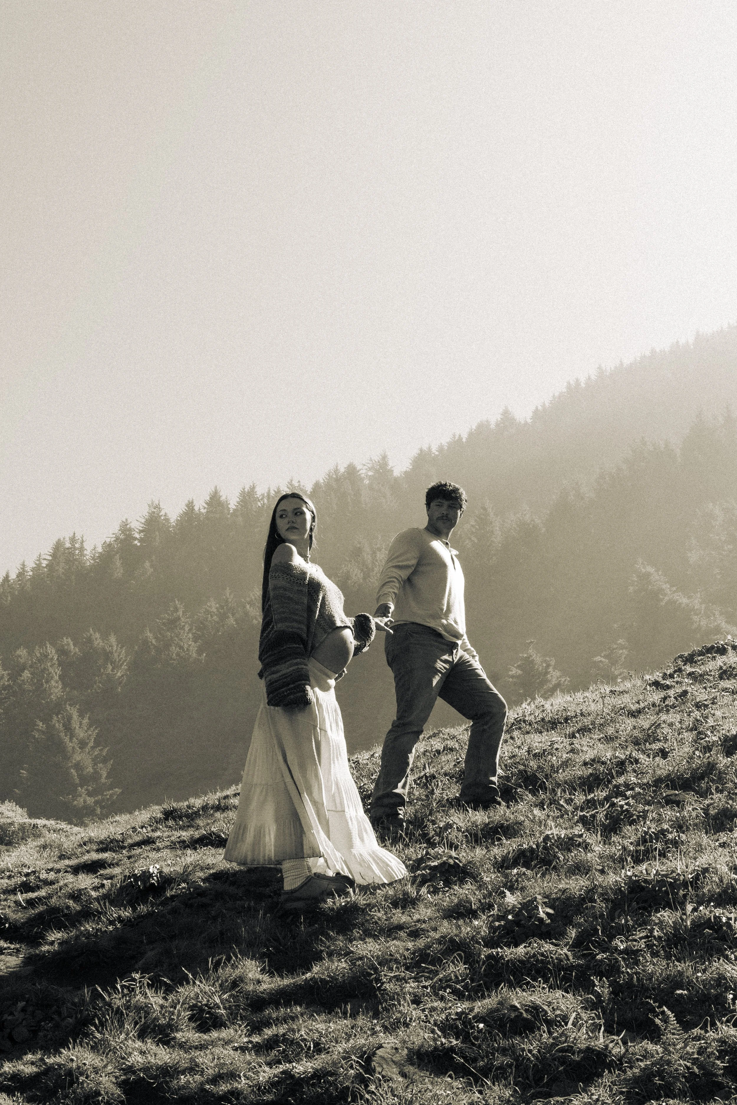 A black and white photo of a young couple holding hands on a grassy hillside with a forested mountain in the background.
