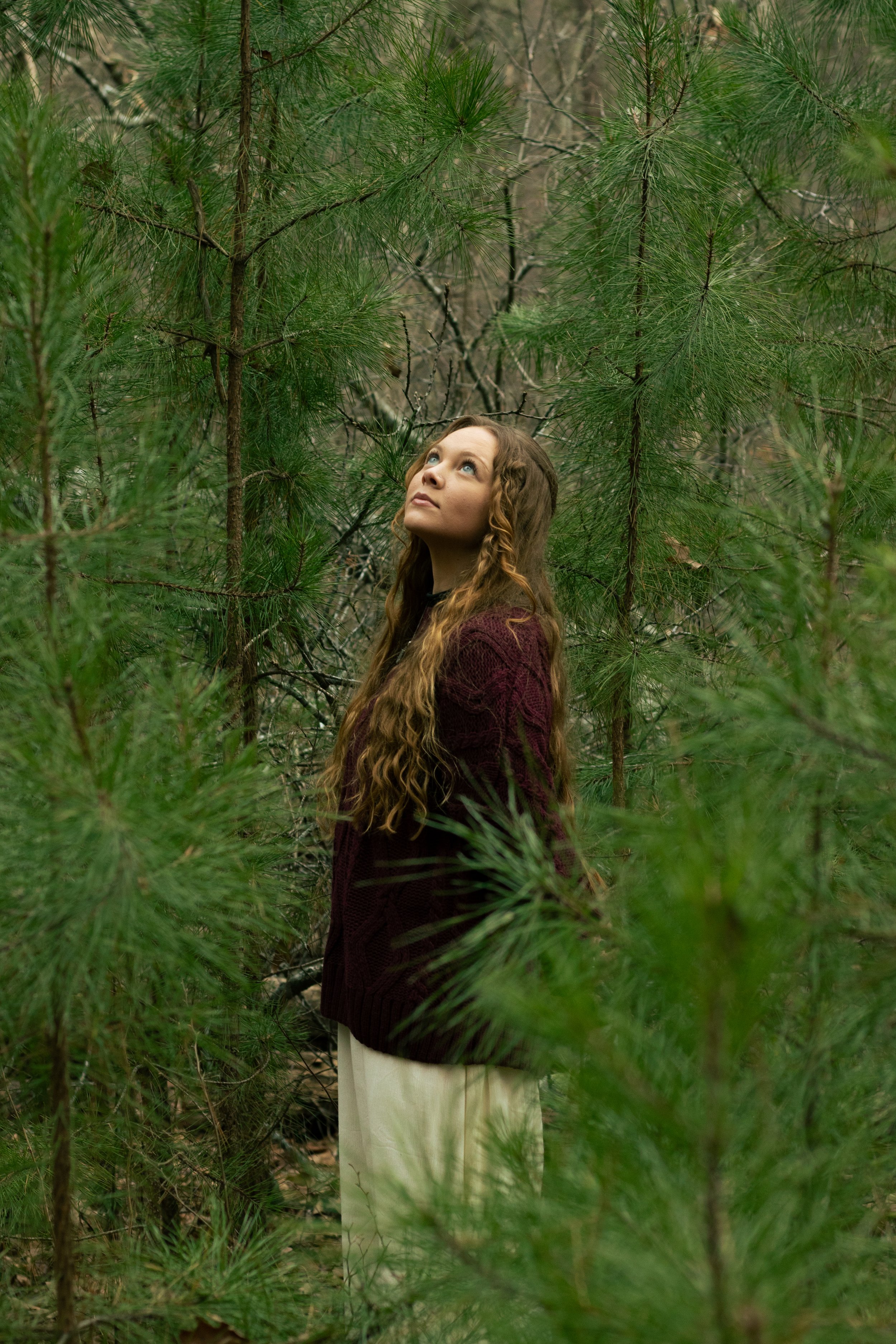 A woman with long, curly hair, wearing a maroon sweater and beige pants, standing amidst green pine trees and looking upward.