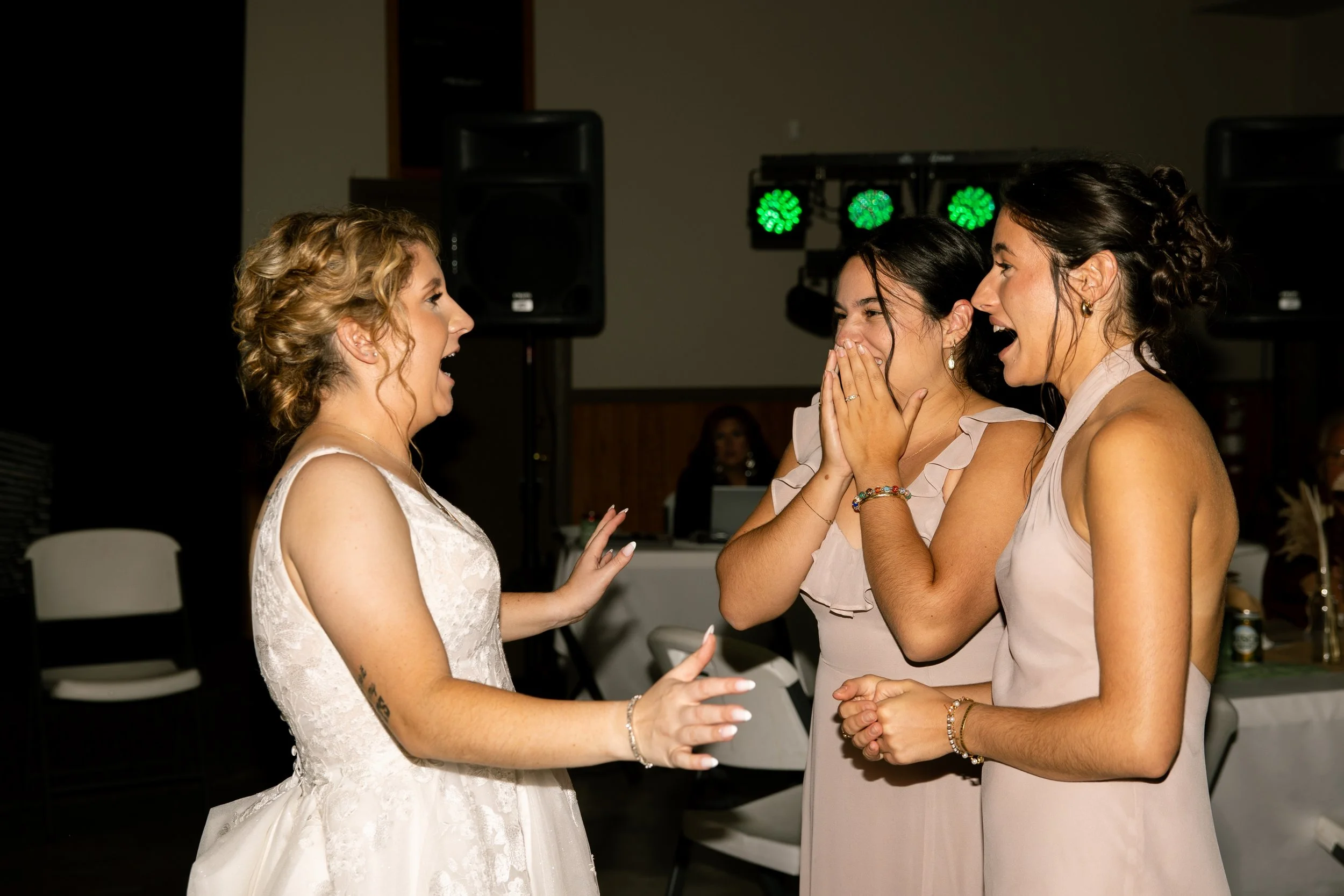 Three women at a wedding reception, one in a white wedding dress and two in beige dresses, look at each other joyfully, laughing and smiling.