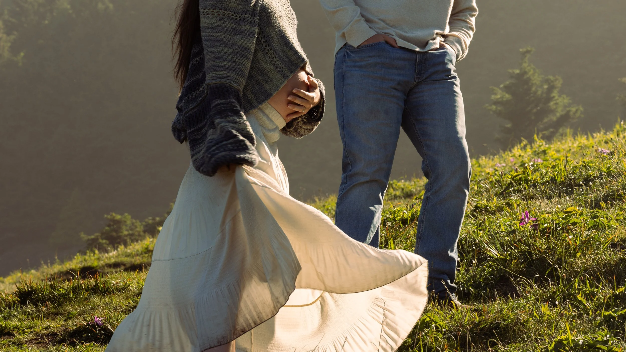 A woman with long hair wearing a knitted sweater, white skirt, and gloves kneels on grass, holding her pregnant belly. Next to her, a man in a gray sweater and jeans stands with his hands in his pockets, on a hillside with green vegetation, pink flow