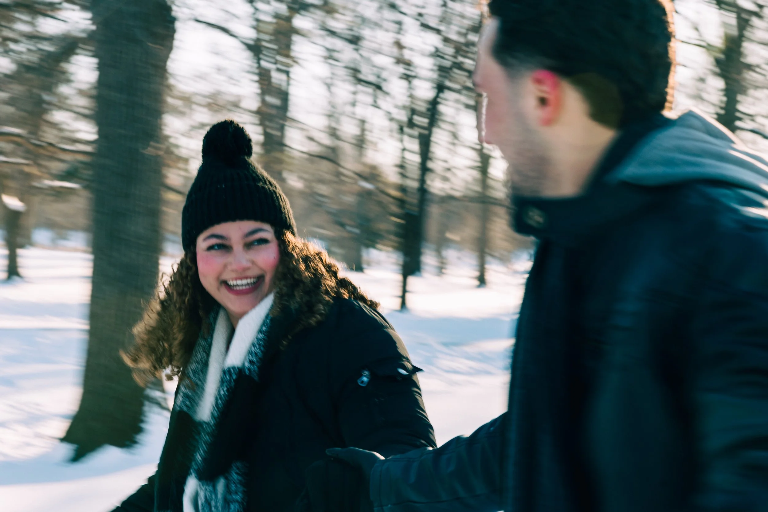 A woman with curly hair, wearing a black knit hat and winter coat, smiling while talking to a man in a black jacket in a snowy forest.