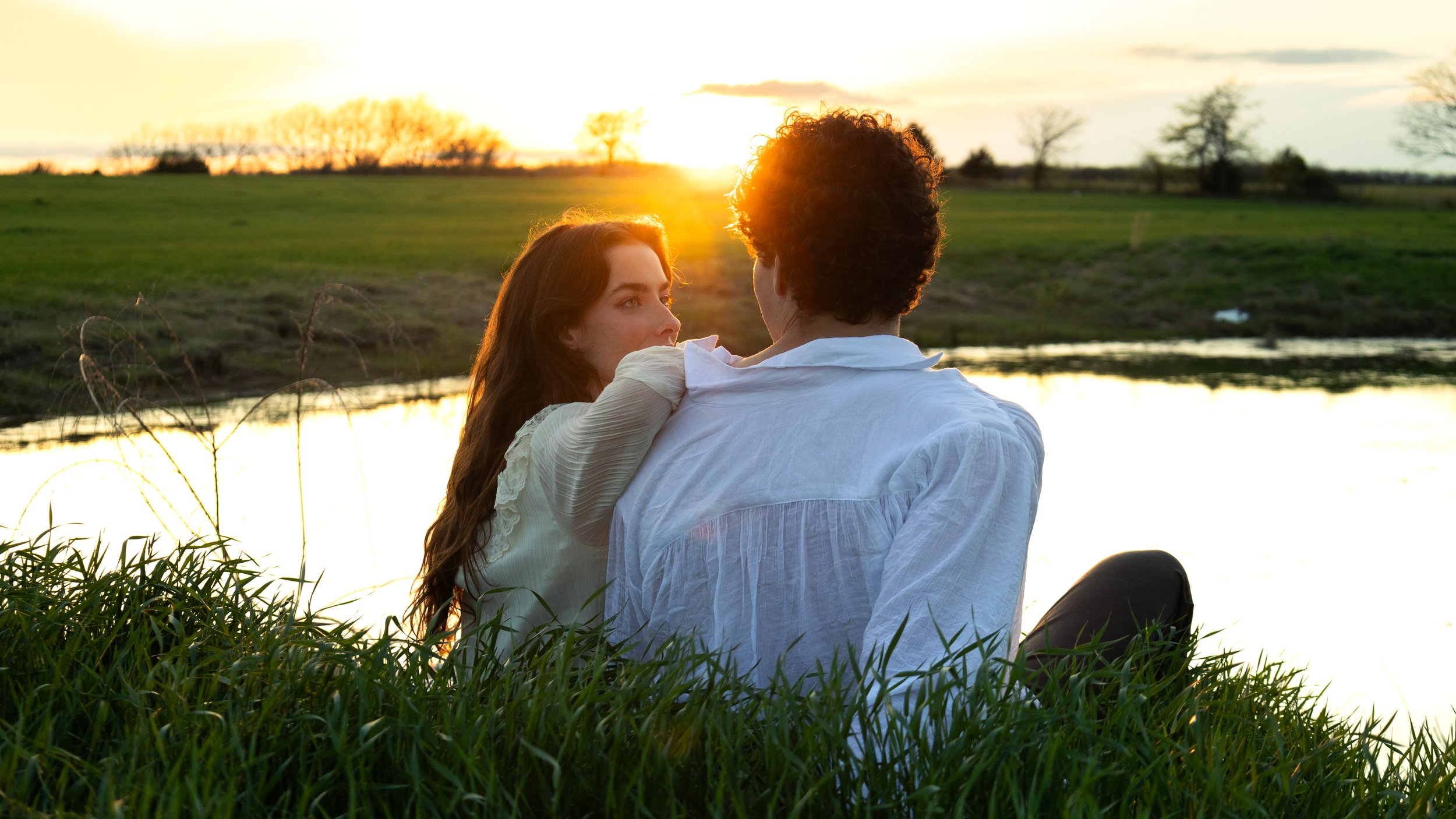 A couple sitting by a lake during sunset, watching each other, with grass in the foreground and trees in the background.