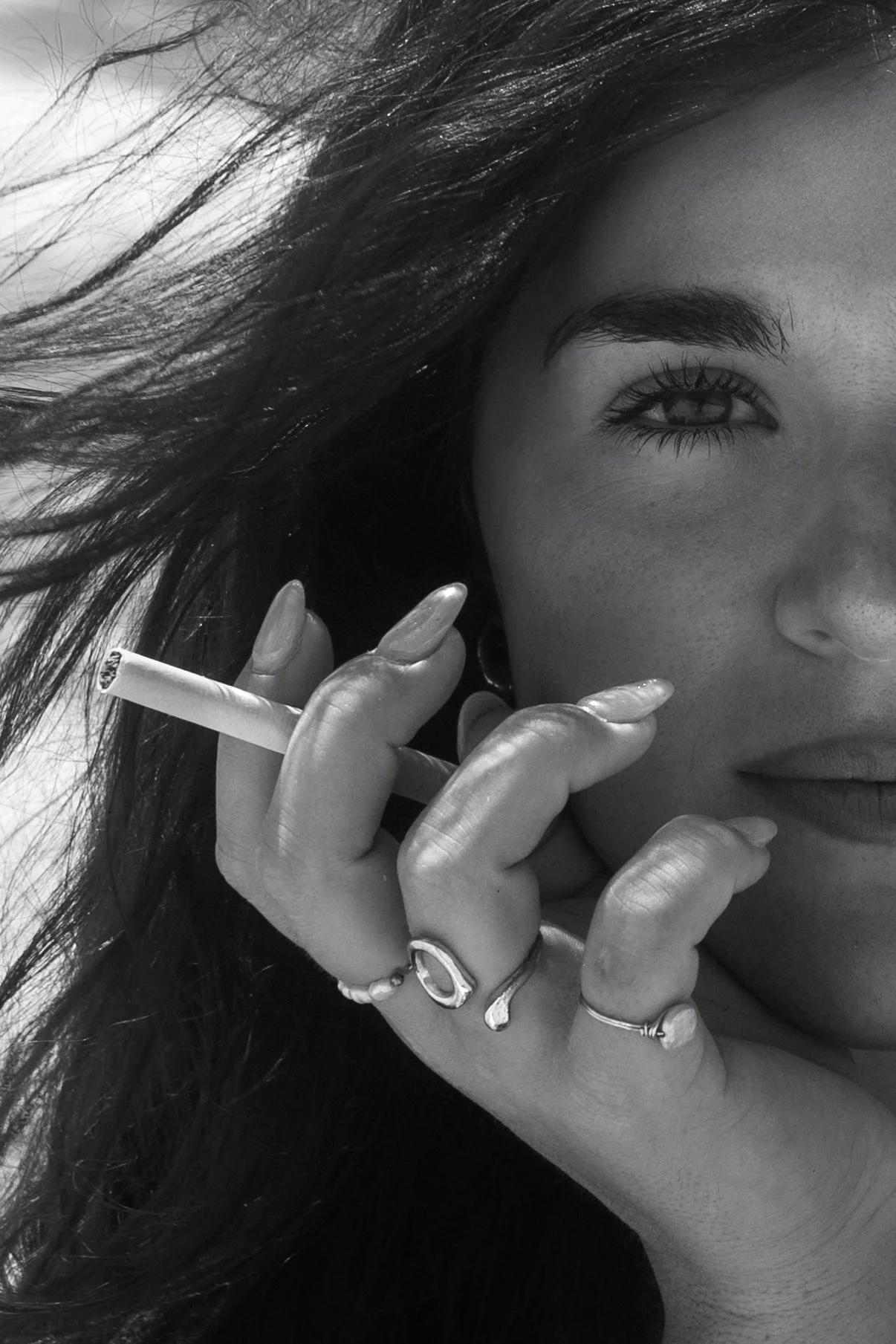 Close-up black and white photograph of a woman with long dark hair, holding a cigarette near her face. The image captures half of her face, emphasizing her eye, eyebrows, and lips. She wears multiple rings on her fingers and has well-manicured nails.