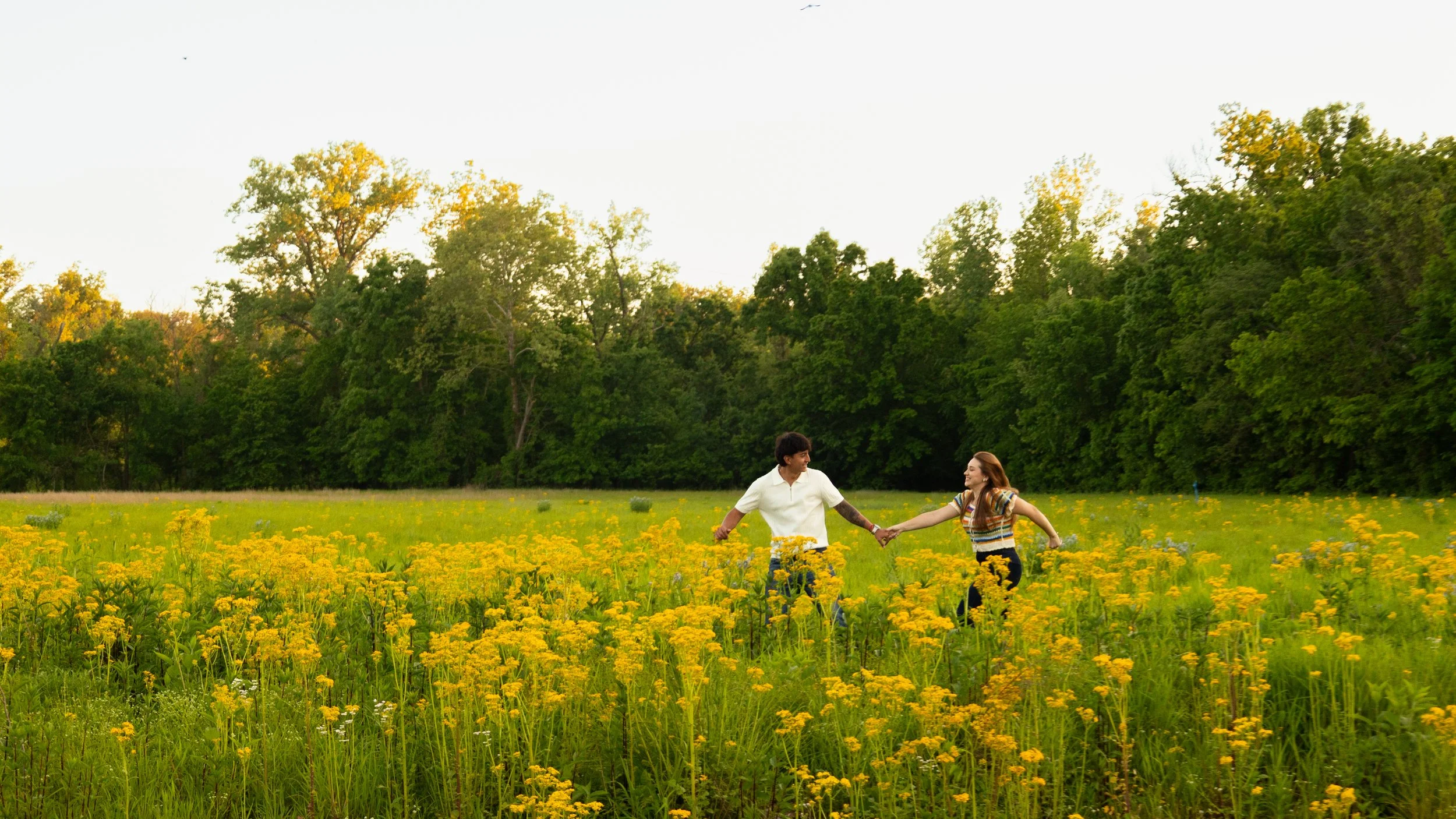A couple holding hands and dancing in a field of yellow flowers with green trees in the background during daytime.