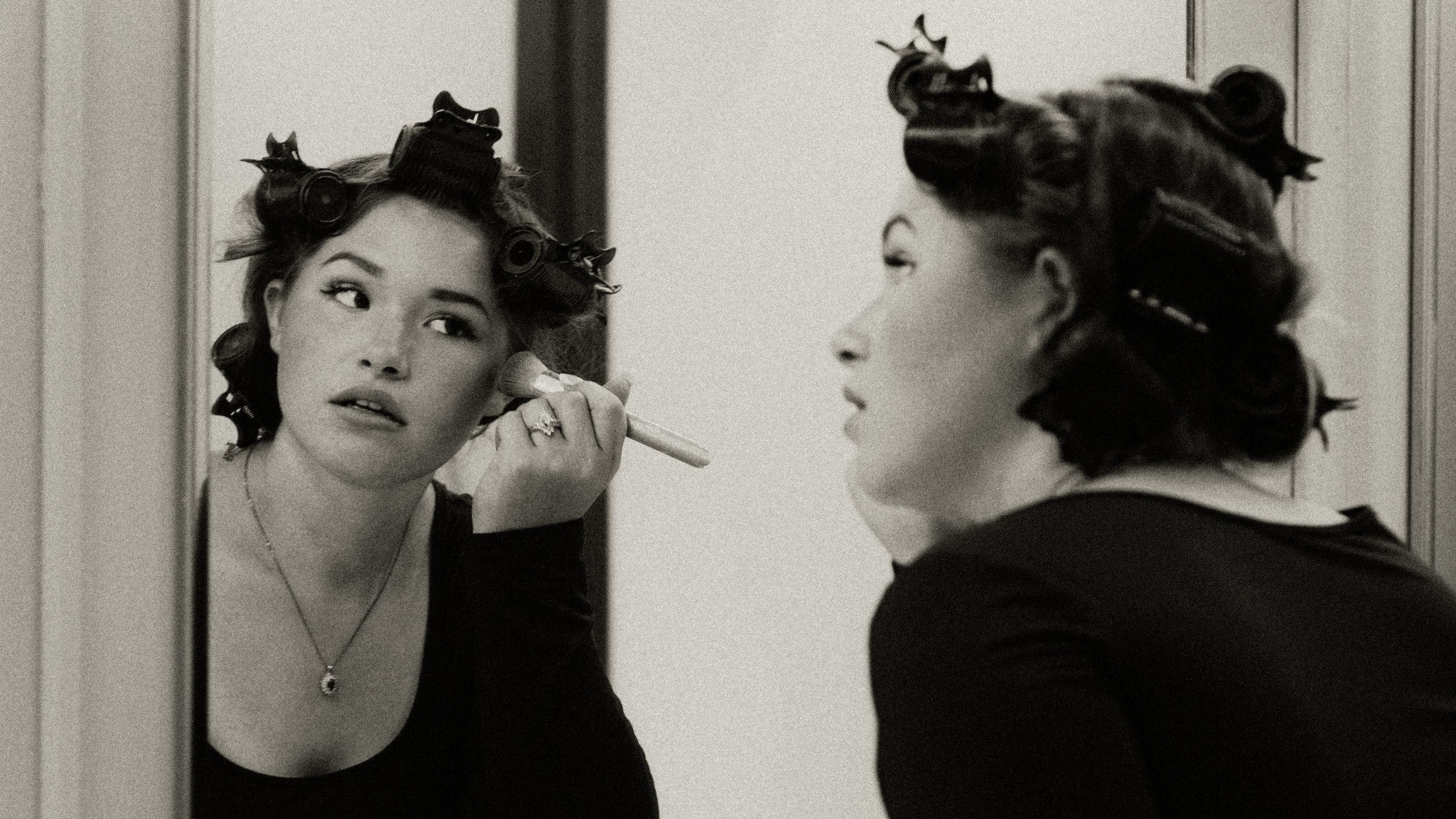 A woman with hair rollers applying makeup in front of a mirror.