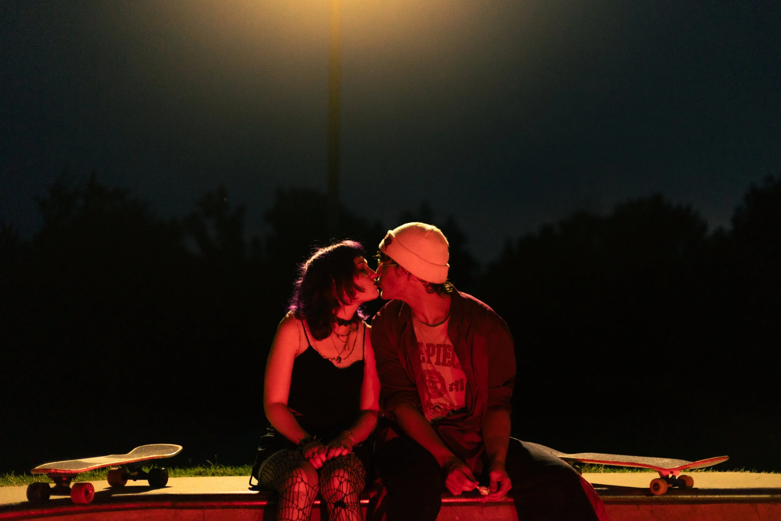 A young woman and a young man are sitting on a bench at night, sharing a kiss under a streetlamp. There are skateboards placed on the ground beside them.