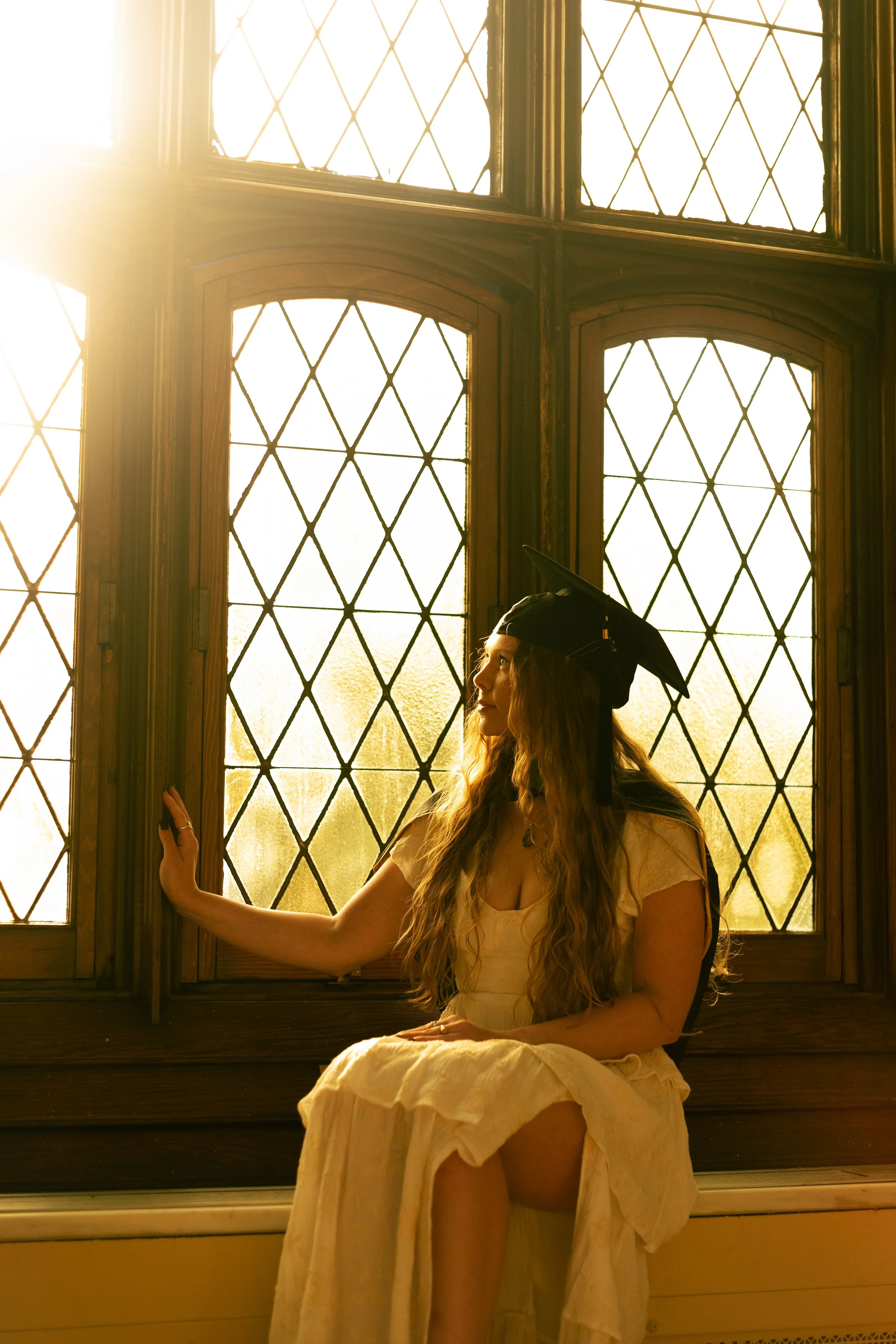 A young woman in a cream-colored dress and graduation cap sitting by stained glass windows with sunlight streaming in.