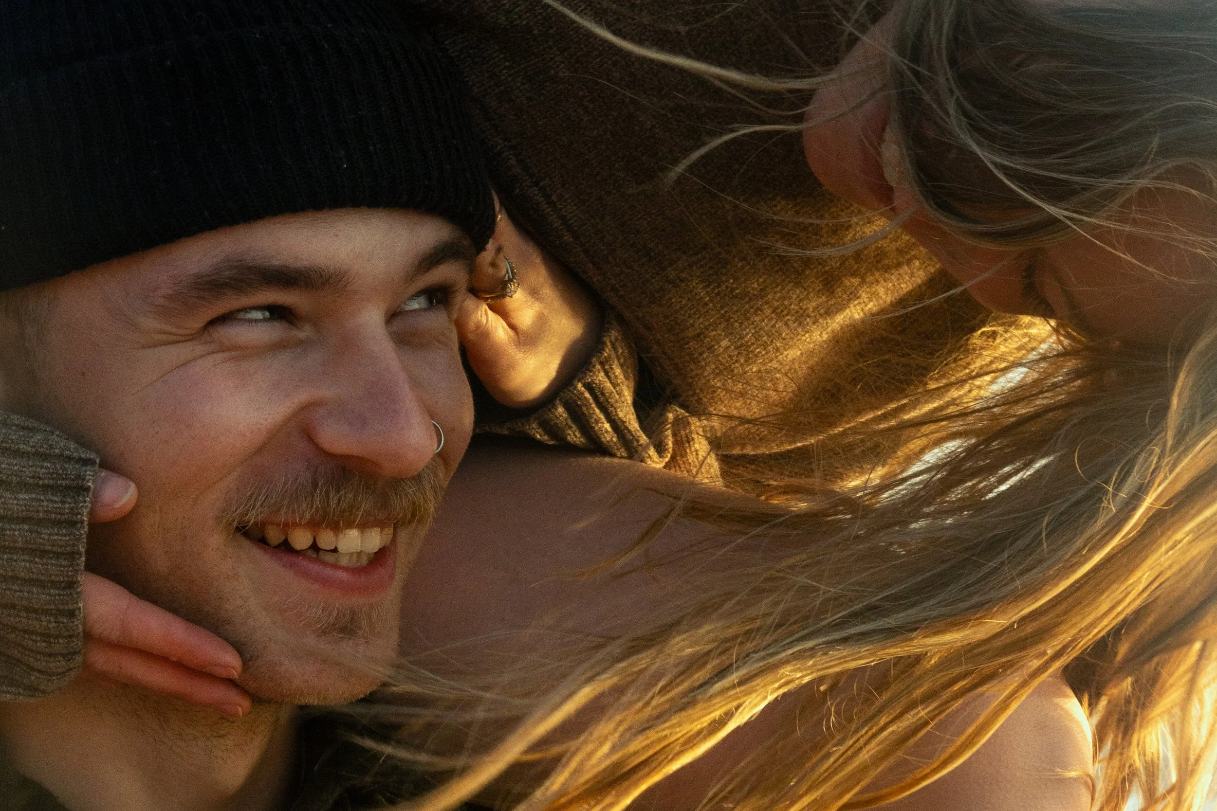 Close-up of a smiling man with a mustache and beard wearing a black beanie, being playfully tangled by a woman with long blonde hair.