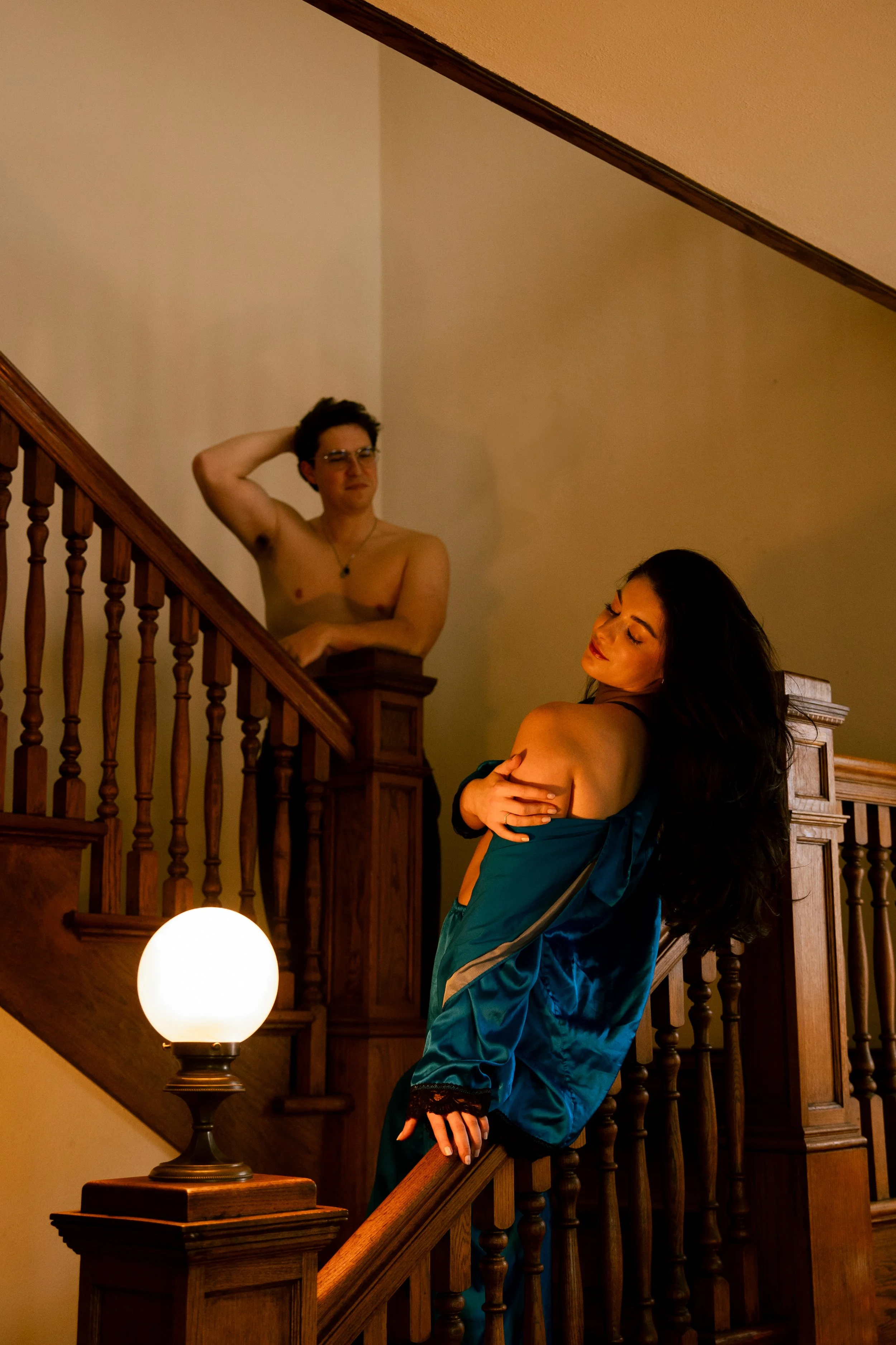 A woman with long dark hair wearing a teal satin robe is standing on a staircase, looking down with her eyes closed and a serene expression. A shirtless man with glasses and short dark hair is leaning on the staircase railing and looking at her, scra