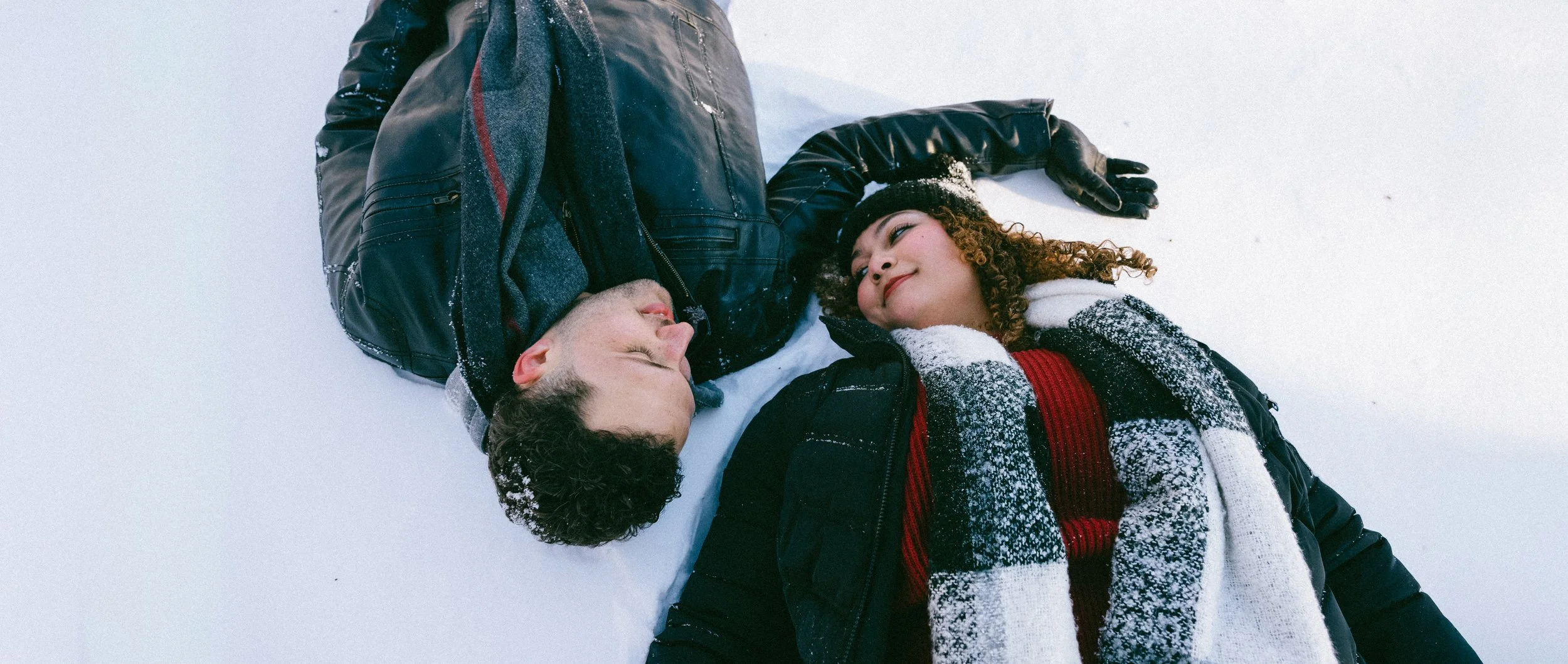 Two people lying on snow, smiling and looking at each other, dressed warmly in winter clothing.