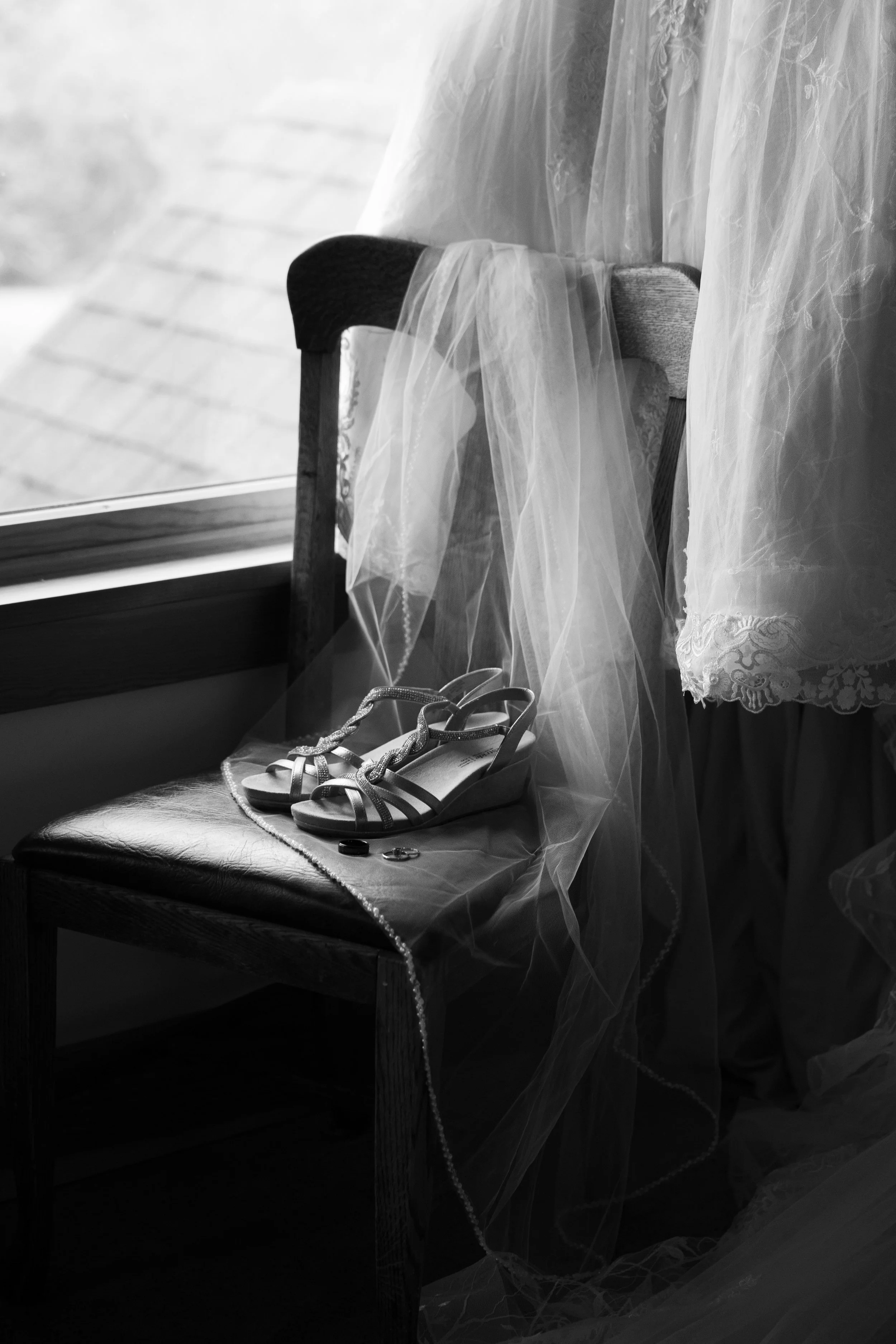 Black and white photo of a chair near a window with wedding shoes, a pearl necklace, and a wedding veil placed on it.