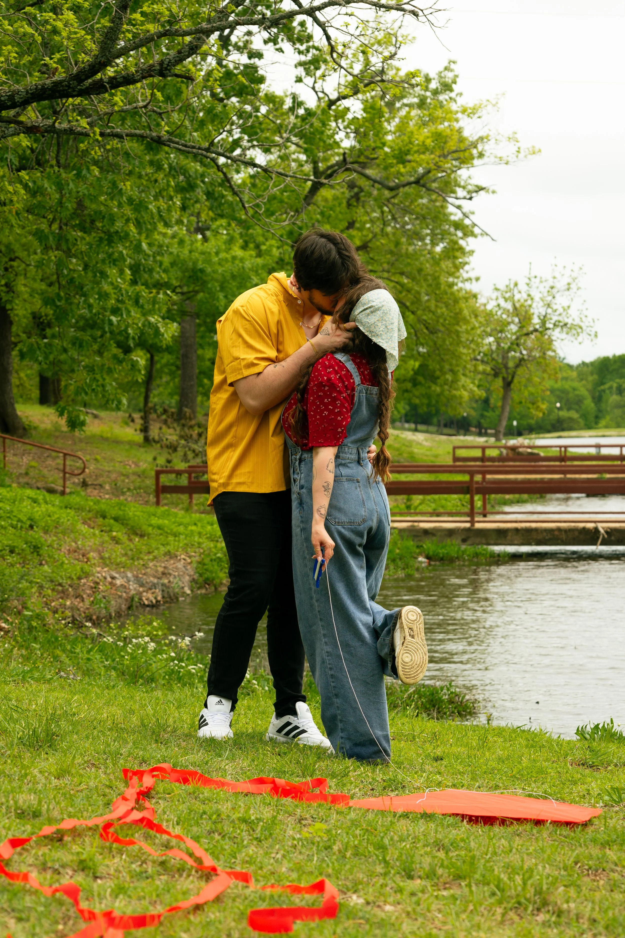 A couple is kissing near a lake in a park, with trees and a bridge in the background. The man wears a yellow shirt and black pants, and the woman has a white headscarf, red blouse, and overalls. There are red ribbons and a small orange parachute on t