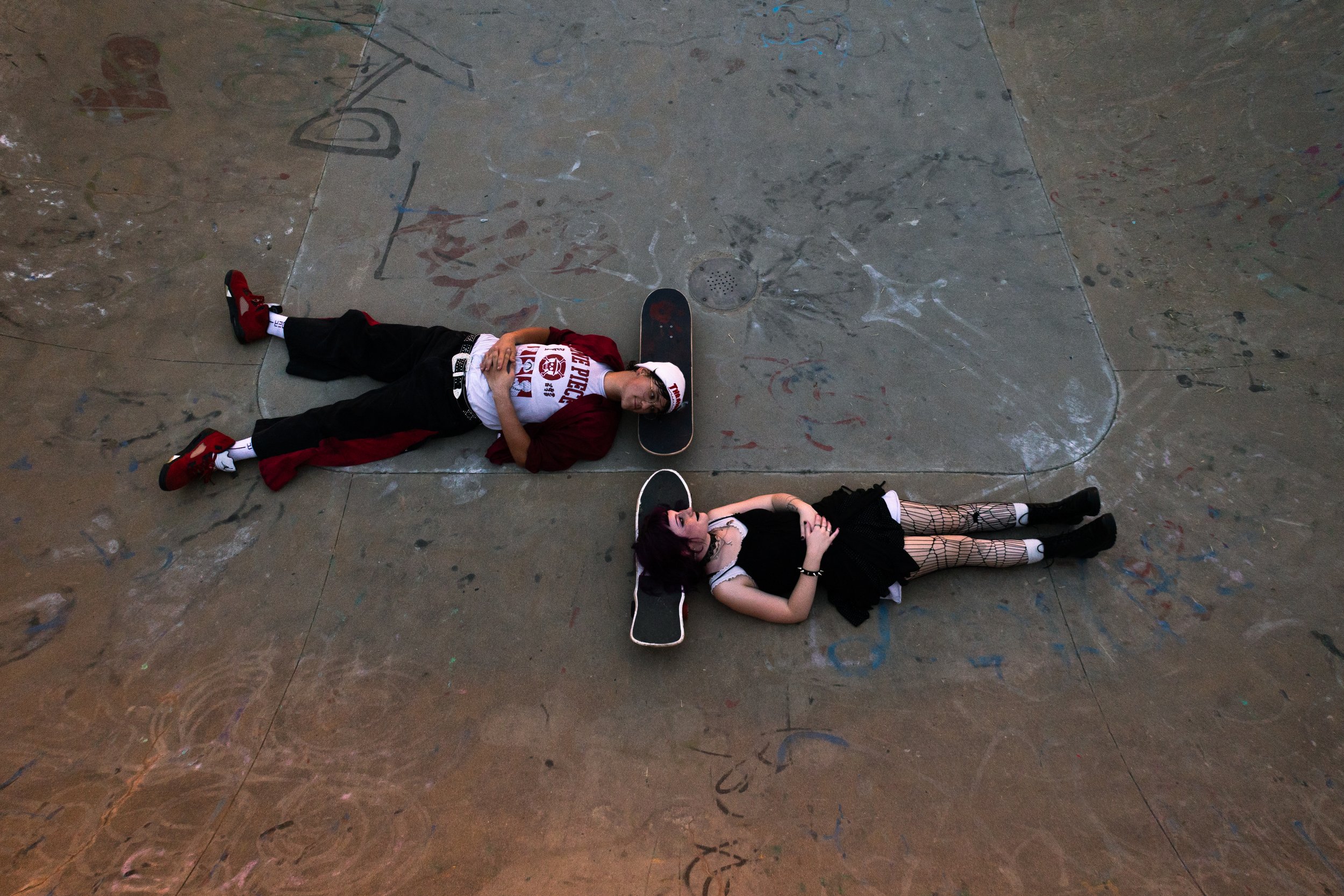 A young man and woman lay on the floor of a skatepark, each with a skateboard underneath their heads, looking relaxed and happy.