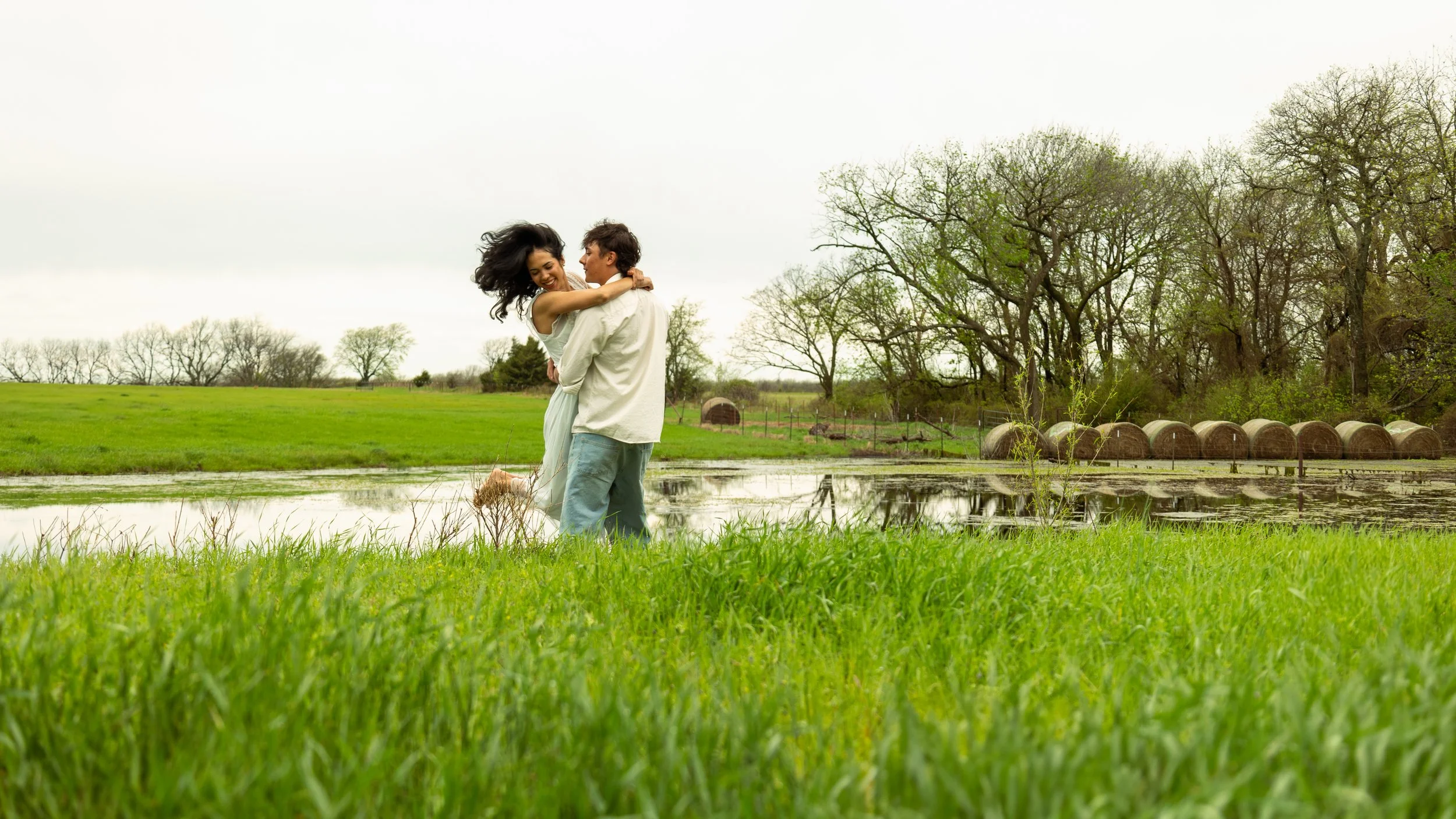 A couple in white clothes embraces and spins near a pond in a green field, with trees and hay bales in the background on an overcast day.