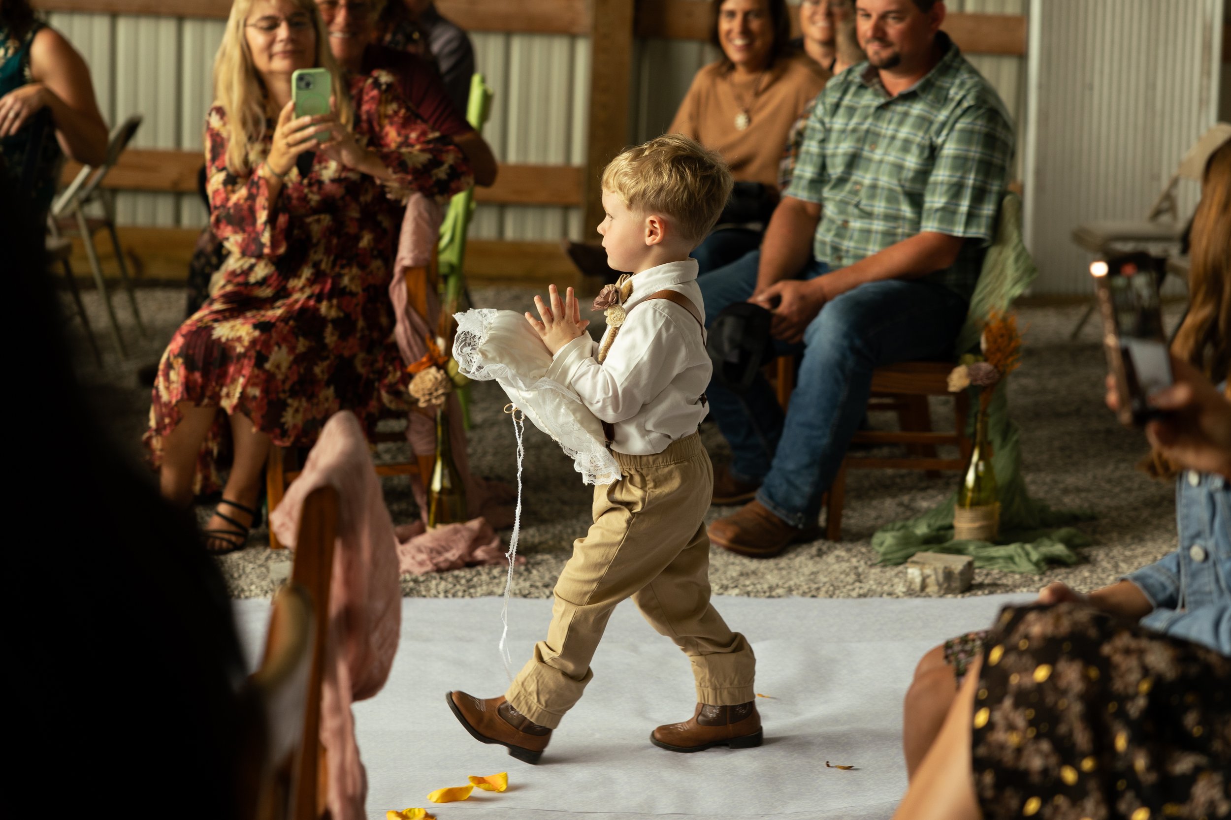 A young boy dressed in vintage-style clothing walking on a white aisle at a wedding or special event, carrying a pillow, with people seated on either side and some taking photos.