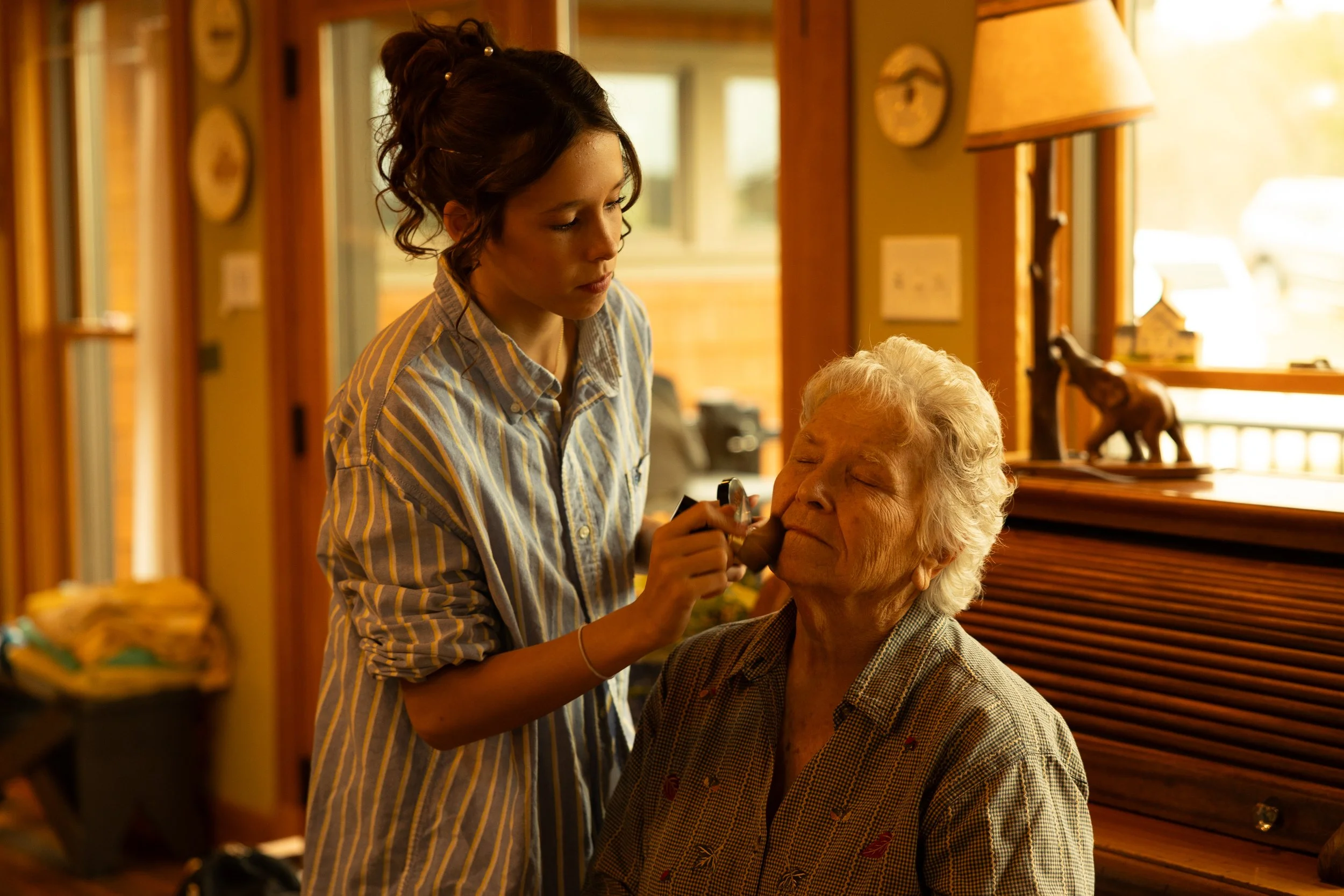 A young woman applying makeup to an elderly woman in a cozy, wood-paneled room with warm lighting, decorative elephant figurines, and a side table.