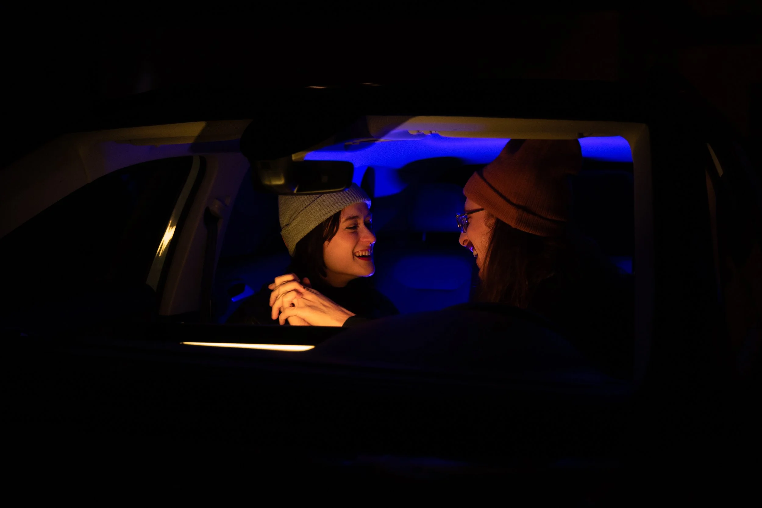 Two women wearing beanies smiling and holding hands inside a car at night, illuminated by blue and orange interior lights.