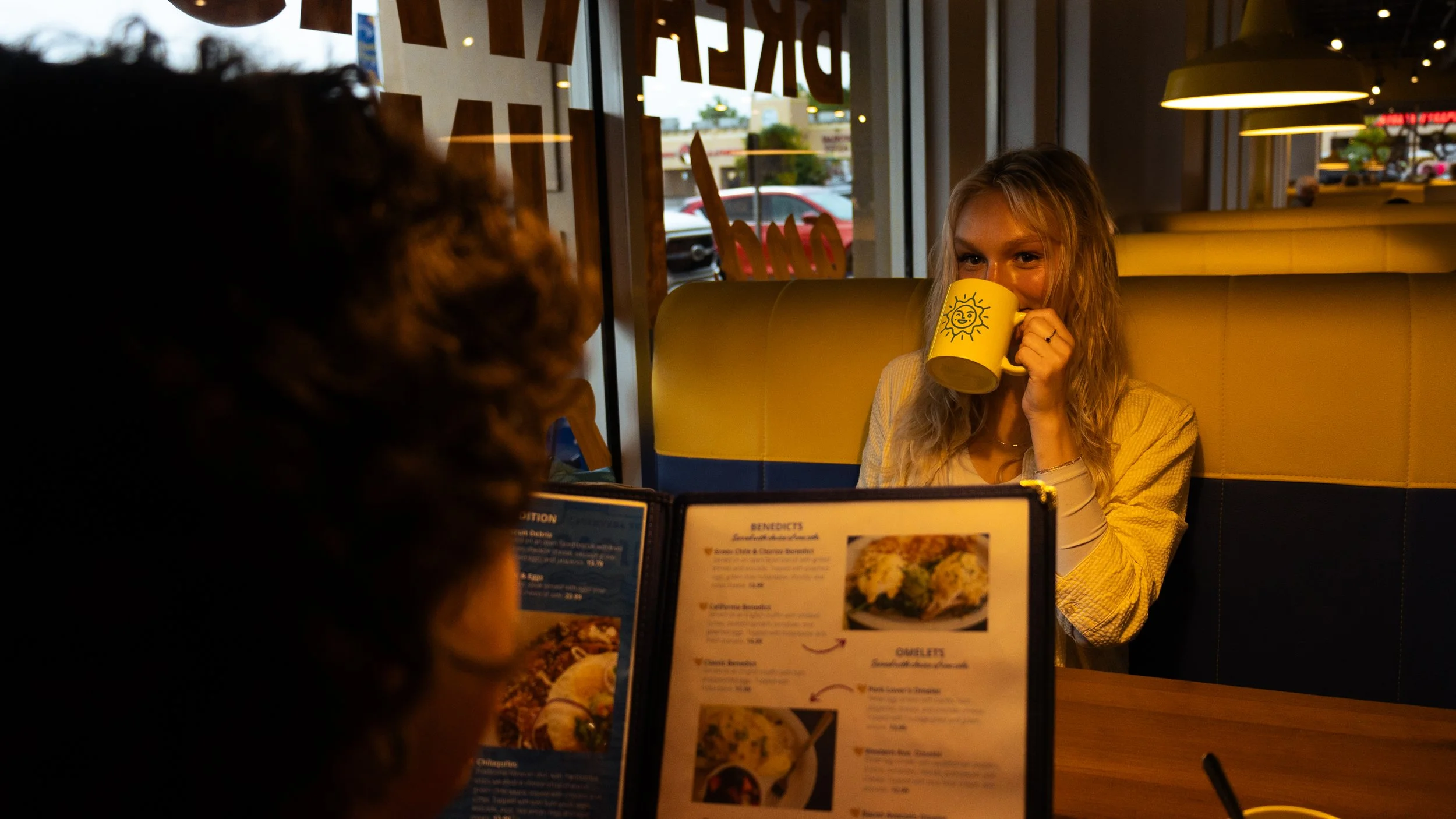 A woman with blonde hair sitting in a restaurant booth, drinking from a yellow mug with a sun drawing, while another person with curly hair looks at her. There is a menu on the table and windows in the background showing a parking lot.