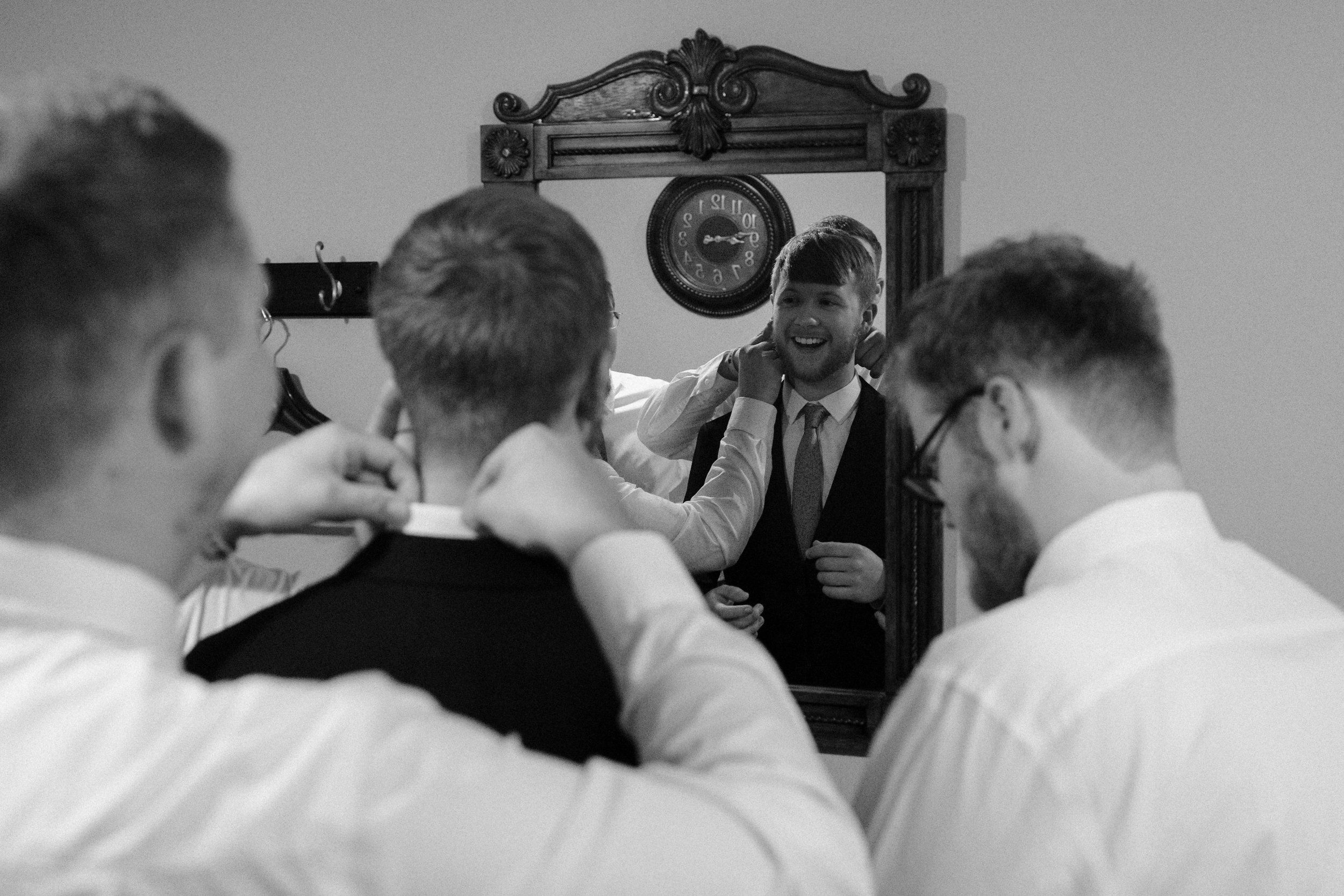 A group of men in formal attire preparing for a wedding, with one man seen in the mirror as he gets dressed.