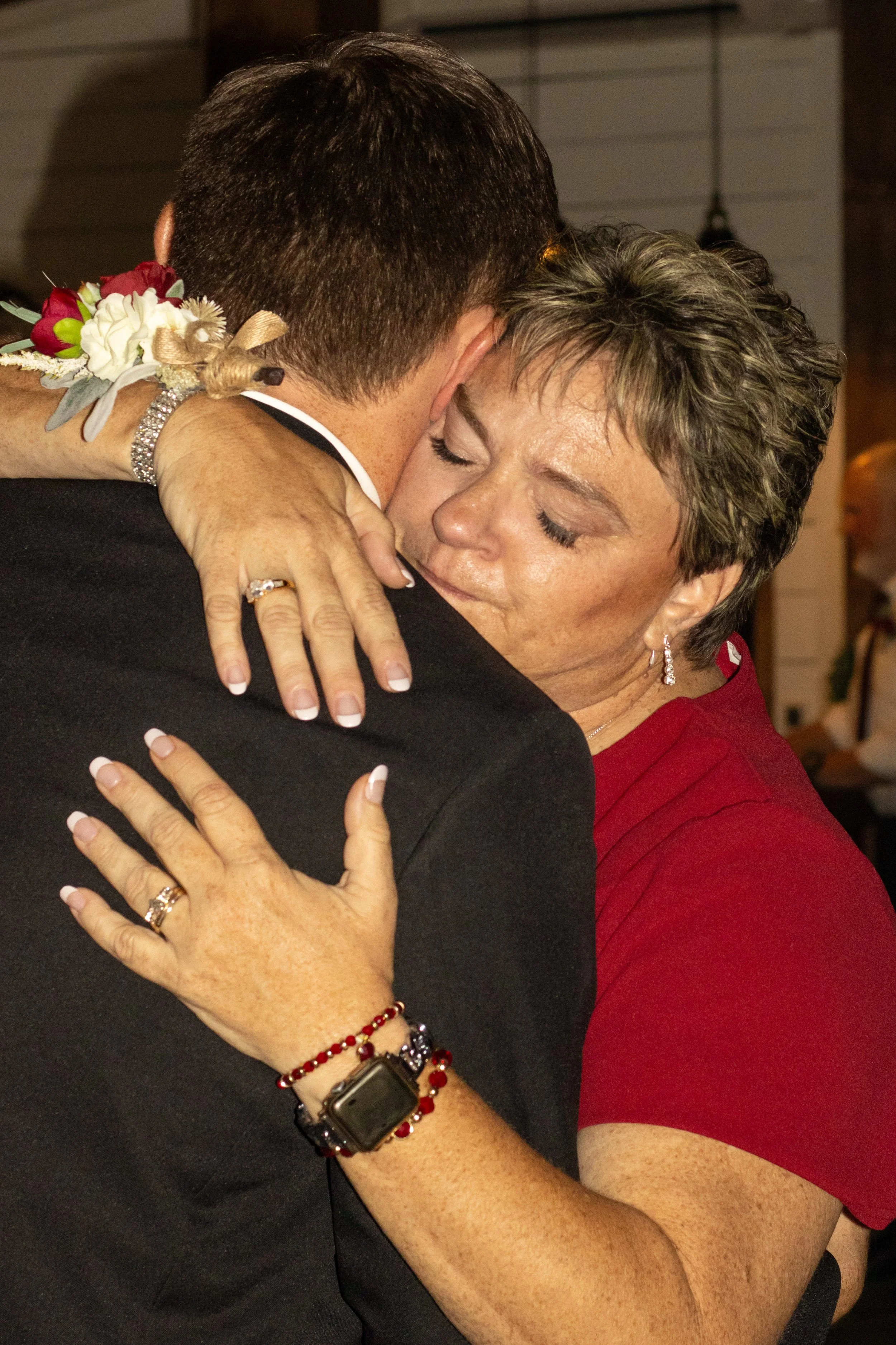 A woman with short gray hair and a woman with short brown hair embrace tightly during a heartfelt moment. The woman has her eyes closed and appears emotional, wearing a red top and jewelry. The woman she is hugging is dressed in a black suit and a bo
