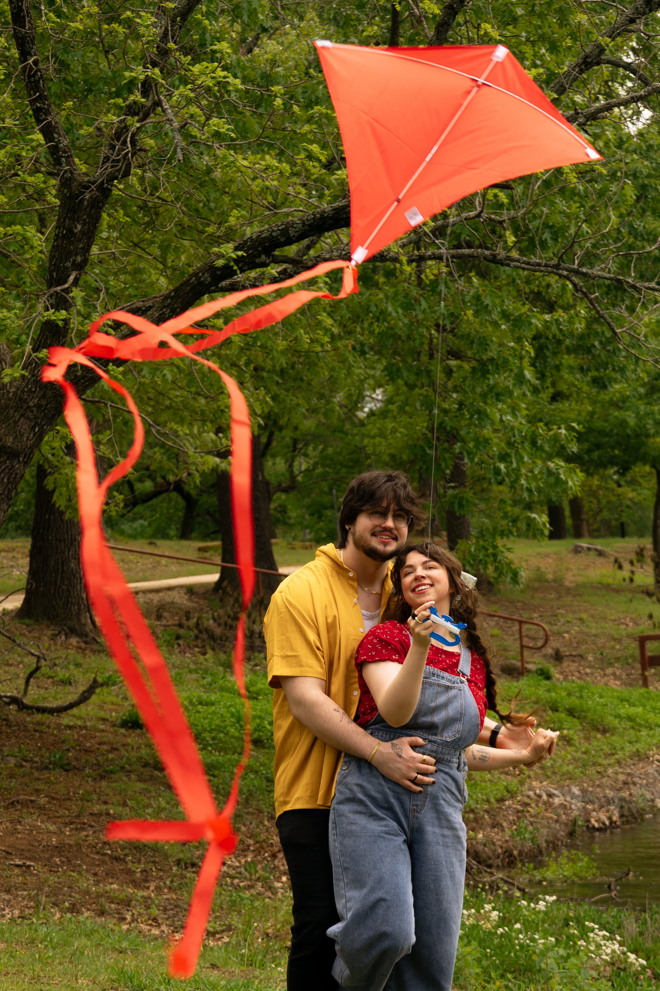 A young couple flying a red kite in a park with green trees, they are smiling and holding a knotted string and a small wind-up toy.