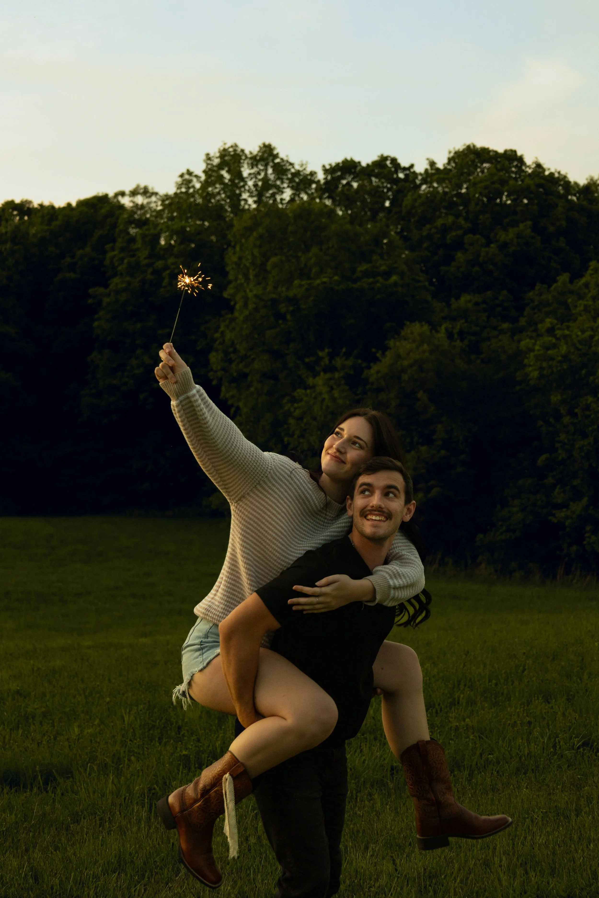 A couple outdoors in a field during sunset, with the woman on the man's back, holding a lit sparkler, both smiling and enjoying the moment.