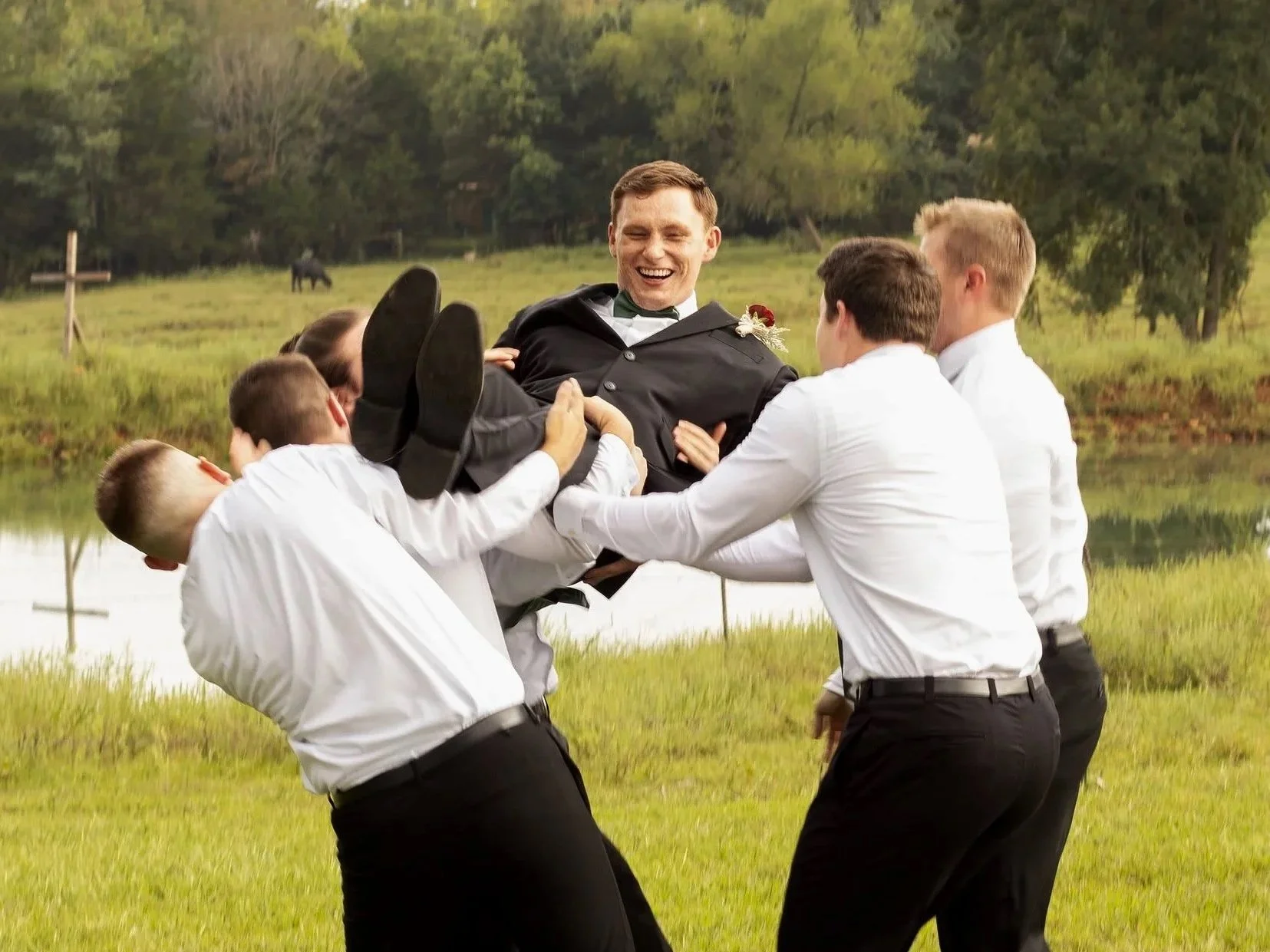 A man in a tuxedo being lifted by five men in white shirts and black pants near a pond, with trees in the background, on a sunny day.